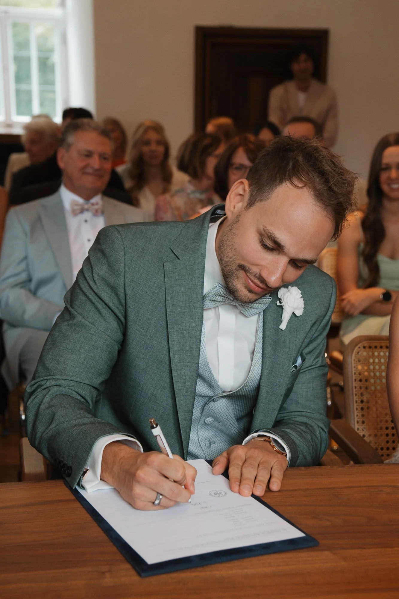 Ein Mann in einem grauen Anzug unterschreibt bei einer Hochzeit, umgeben von Gästen im Raum.