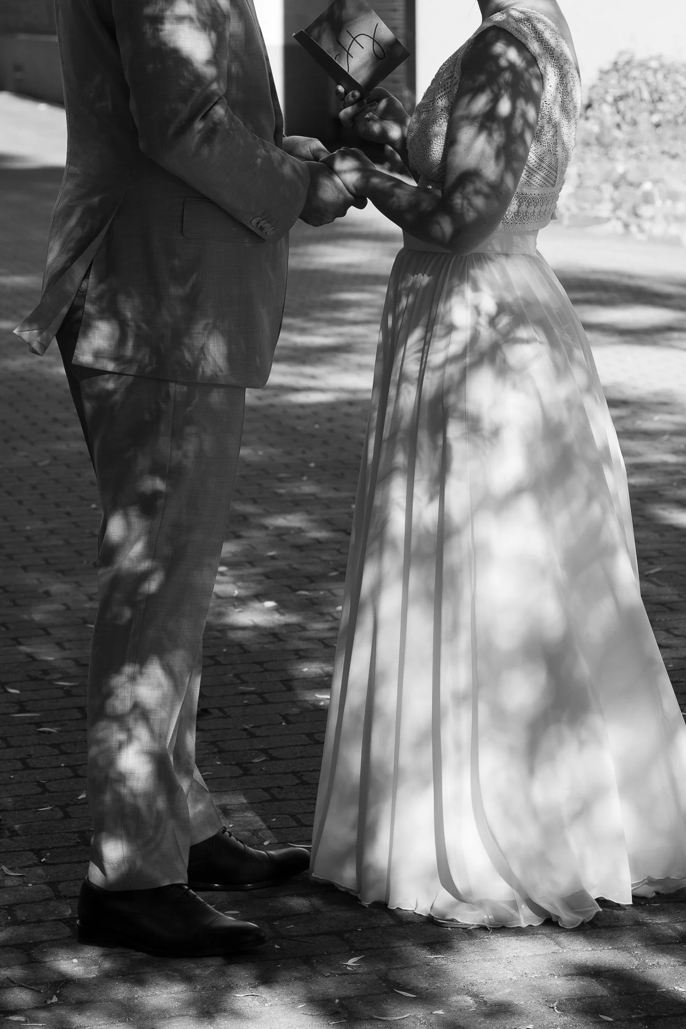 Ein Brautpaar bei einer standesamtlichen Hochzeit, sie hält einen Ehering in der Hand, draußen auf gepflastertem Boden, mit Schatten von Blättern auf ihnen.
