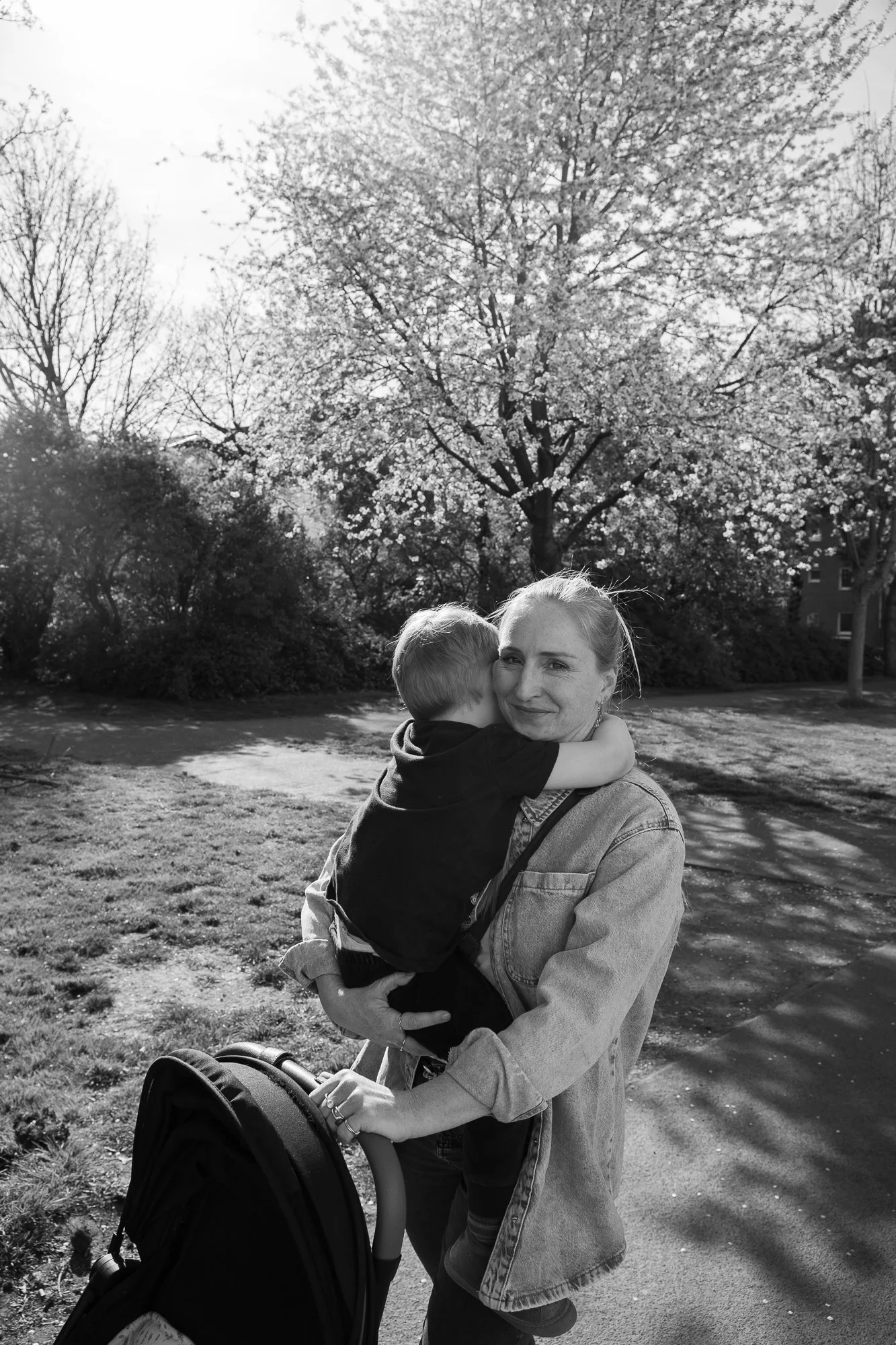 Eine Frau hält einen Jungen, der ihn küssend umarmt, während sie einen Kinderwagen schiebt, im Freien mit Bäumen im Hintergrund.