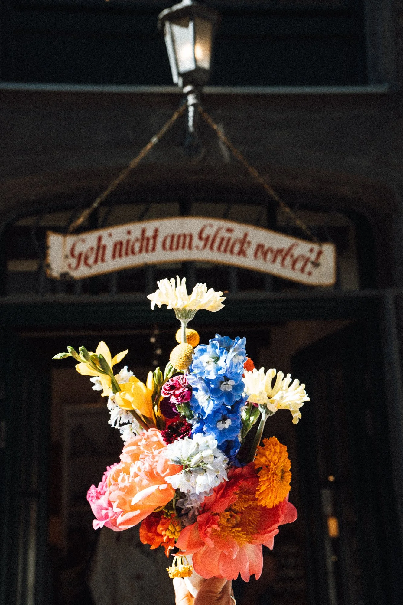 Bunter Blumenstrauß vor dunklem Gebäude mit hängendem Schild, das auf Deutsch sagt: 'Geh nicht am Glück vorbei'.