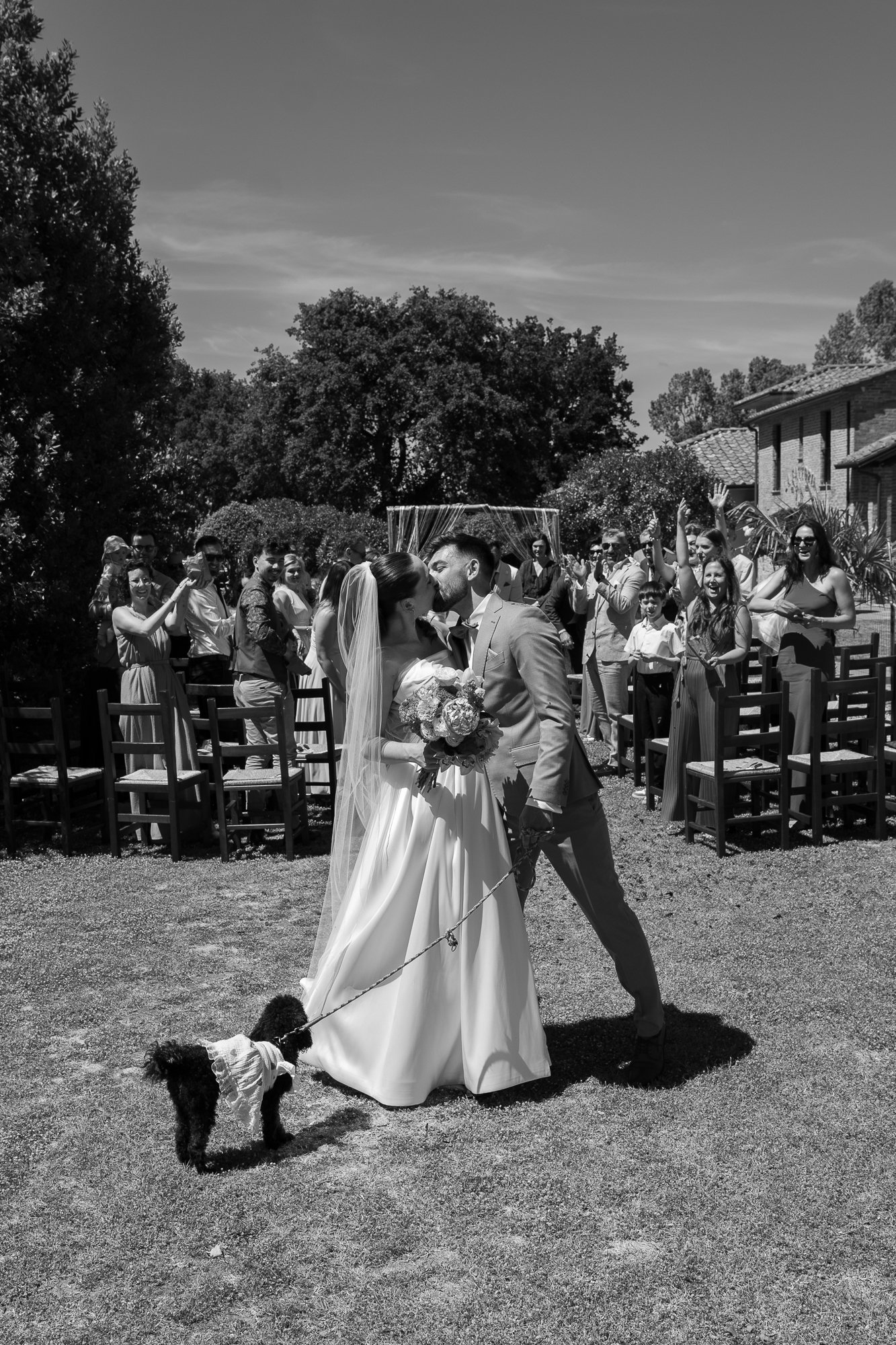 Braut und Bräutigam küssen sich bei einer Hochzeit im Freien, umgeben von lachenden Hochzeitsgästen, mit einem kleinen Hund an der Leine auf dem Rasen, alles in Schwarzweiß.
