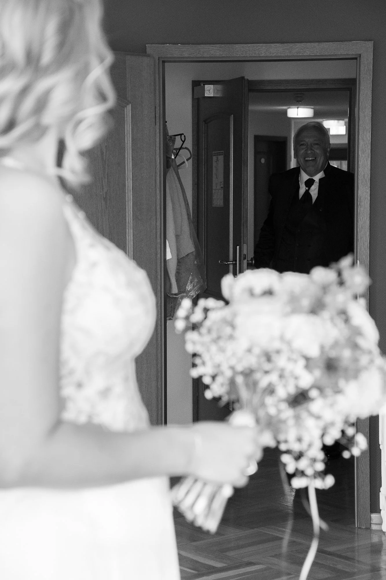 Ein Mann begrüßt eine Braut mit einem Lächeln, im Vordergrund eine Frau mit Brautkleid und Blumenstrauß.