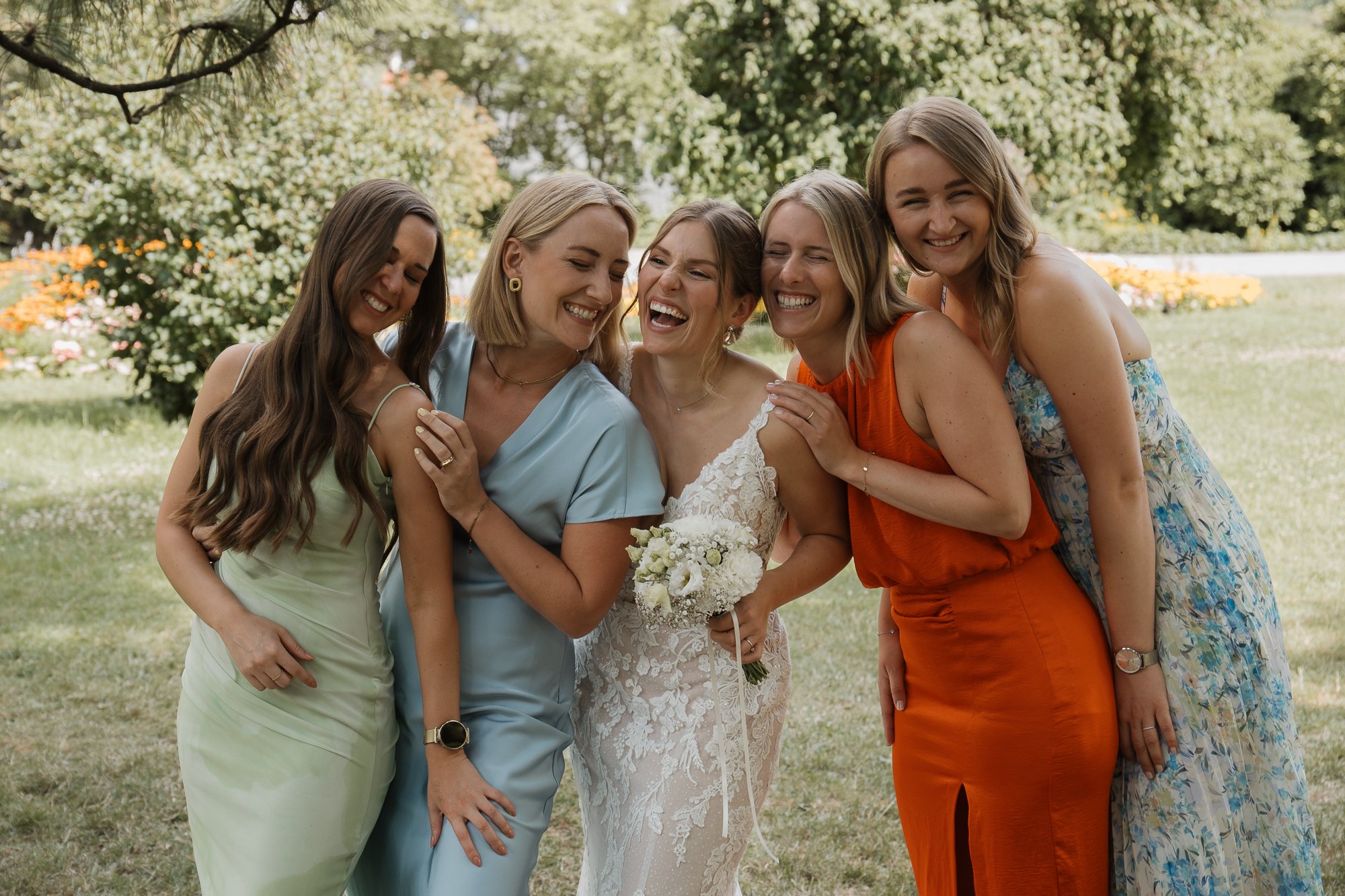 Fünf Frauen beim Lachen im Garten, eine davon ist Braut in weißem Kleid, die anderen sind Freundinnen in farbigen Kleidern.