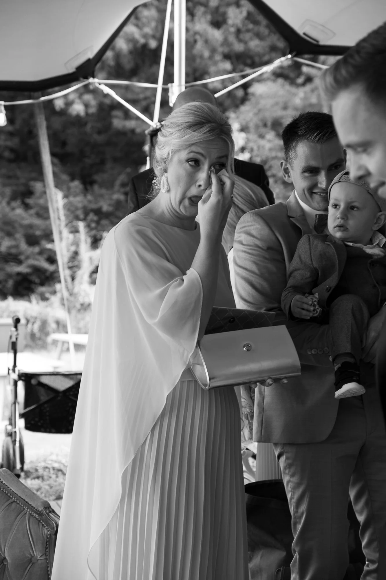 Eine trauernde Frau bei einer Hochzeit, die weint, während sie eine Träne wegwischt, mit einer Gruppe von Menschen im Hintergrund, im Freien, bei einer Zeremonie.