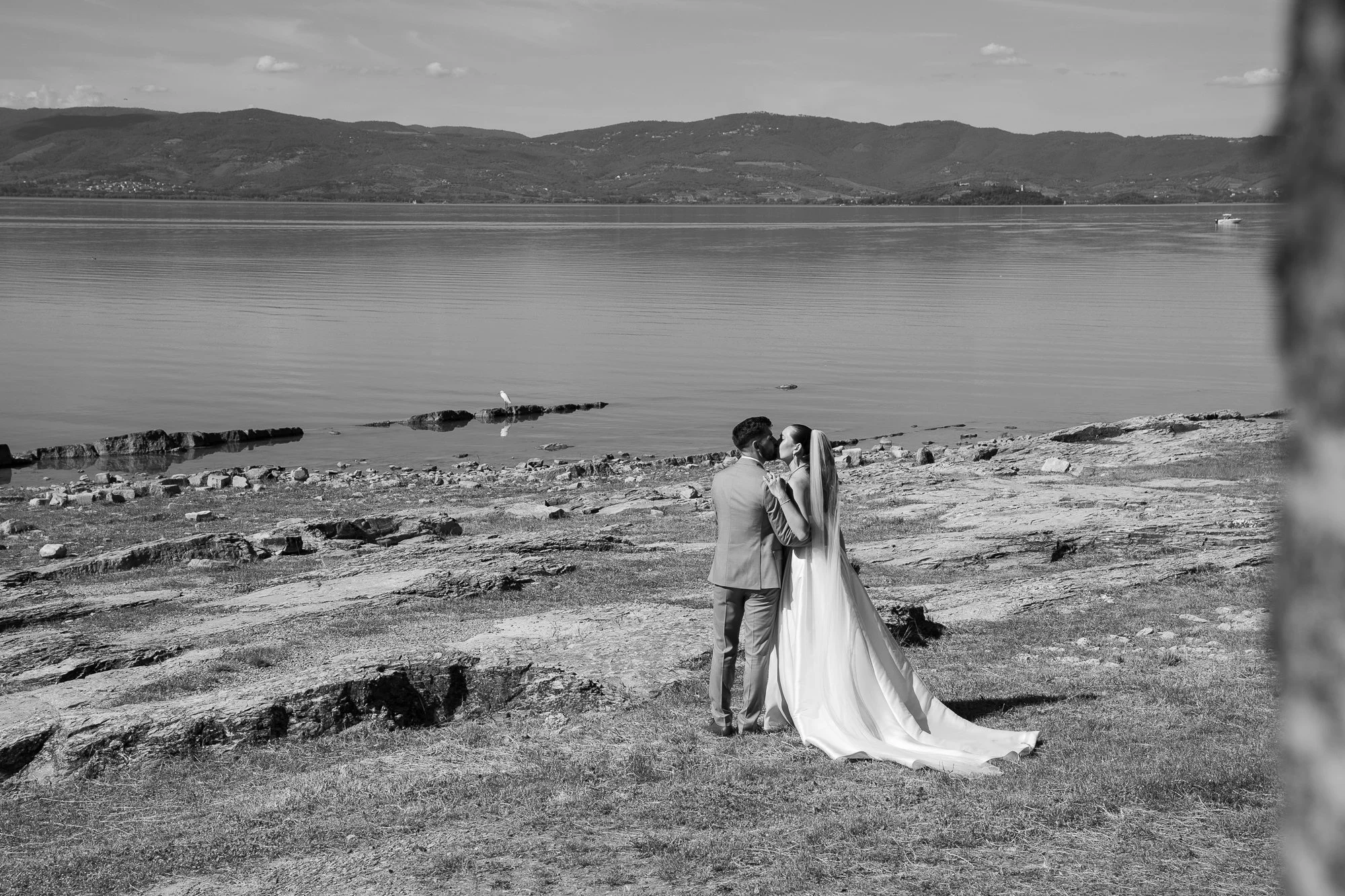 Ein Brautpaar küsst sich am Seeufer, im Hintergrund Wasser und Berge, Schwarz-Weiß-Fotografie.