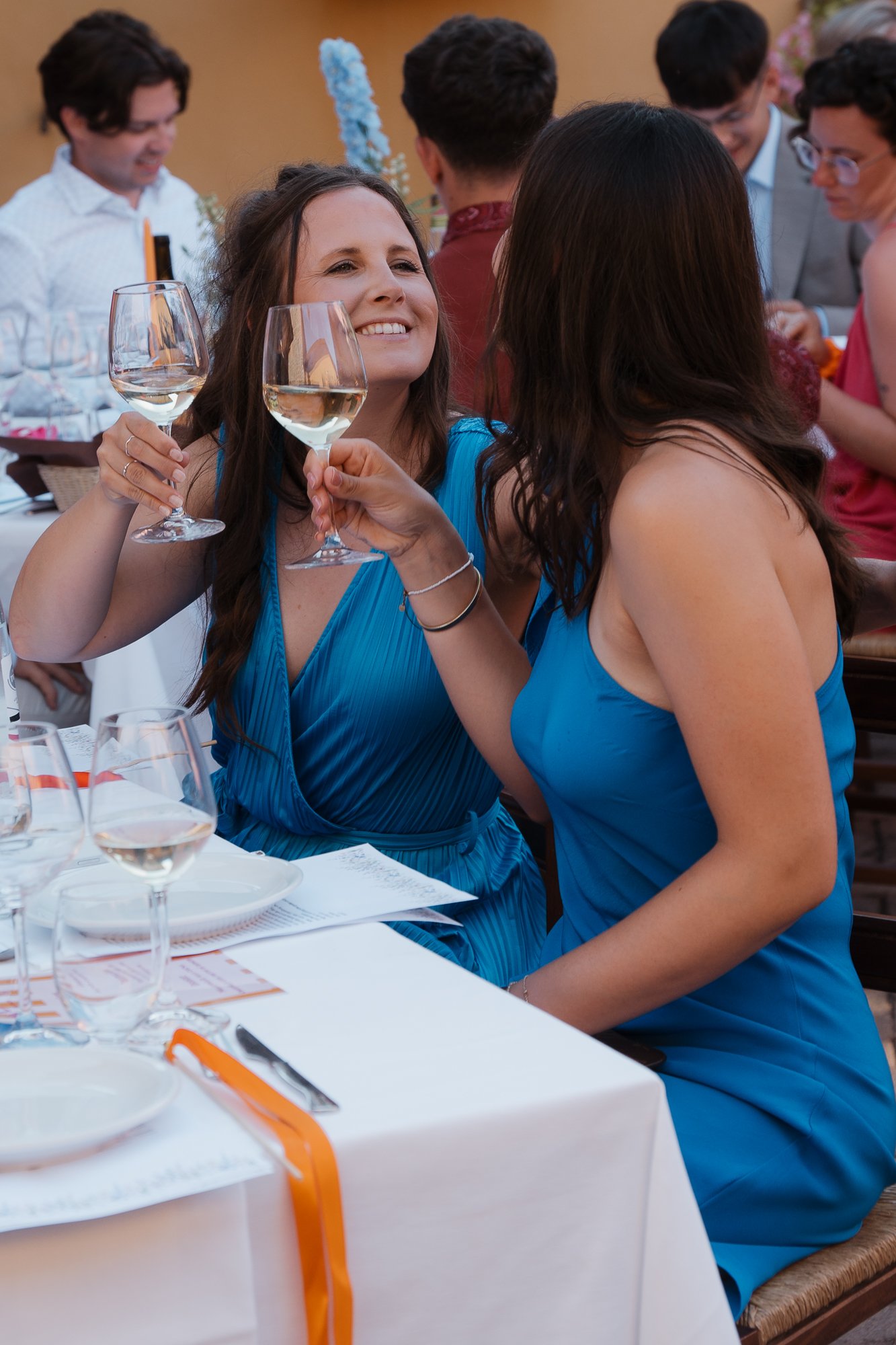 Zwei Frauen in blauen Kleidern stoßen bei einer Feier mit Weißweingläsern an, während im Hintergrund weitere Gäste an einem festlichen Abendessen teilnehmen.