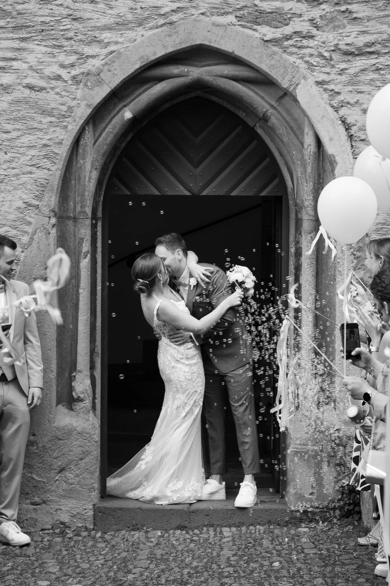 Ein Brautpaar versinkt sich in Kuss, umgeben von Freundinnen und Freunden, bei einer Hochzeit vor einem historischen Steingebäude, mit Partygästen, die Luftballons und Seifenblasen halten.
