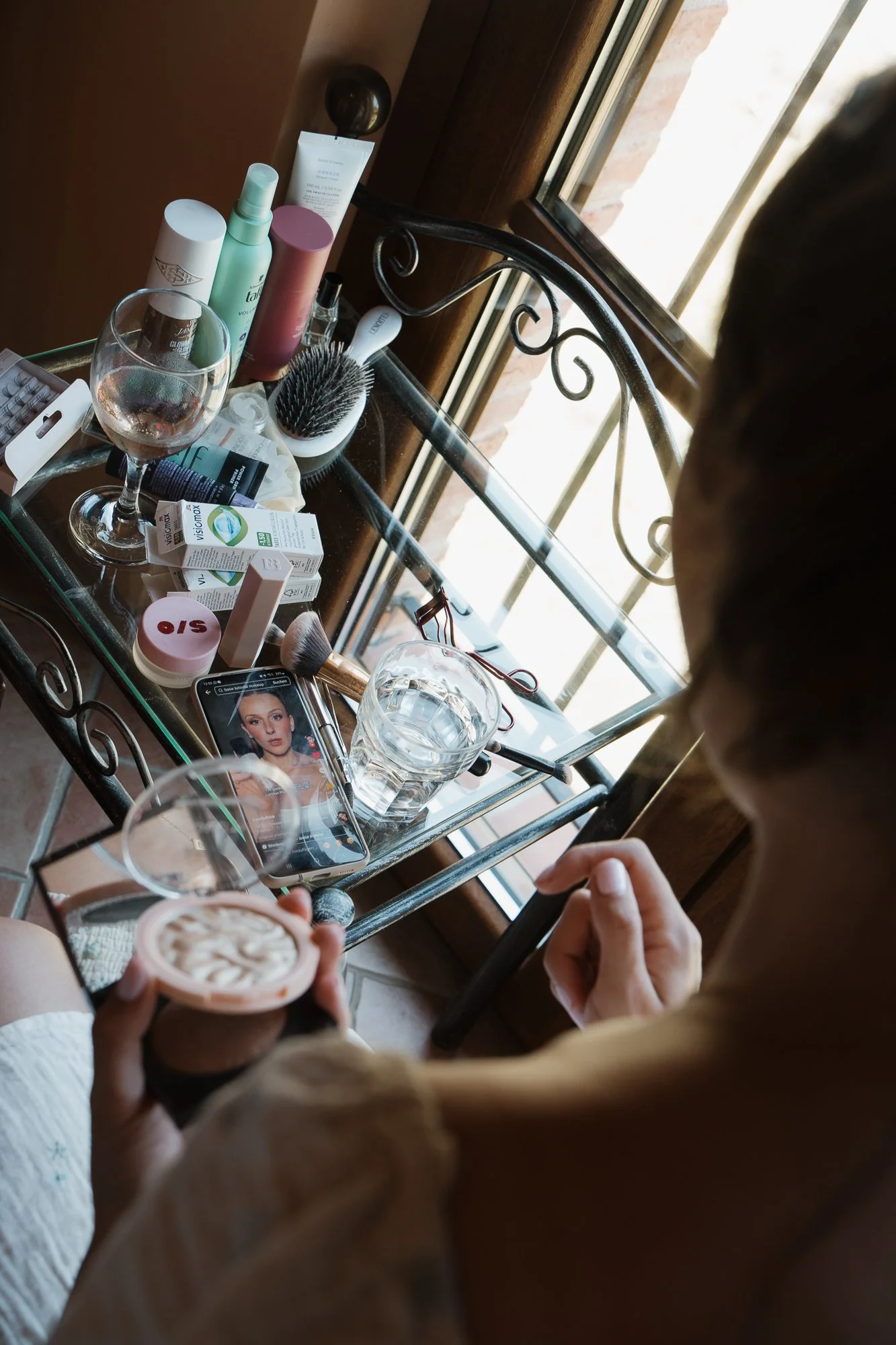 Nahaufnahme eines Schminktisches mit Make-up-Produkten, einem Smartphone mit einem Beauty-Video, einem Trinkglas Wasser, und einer Person, die eine Creme hält. Der Tisch steht vor einem Fenster.