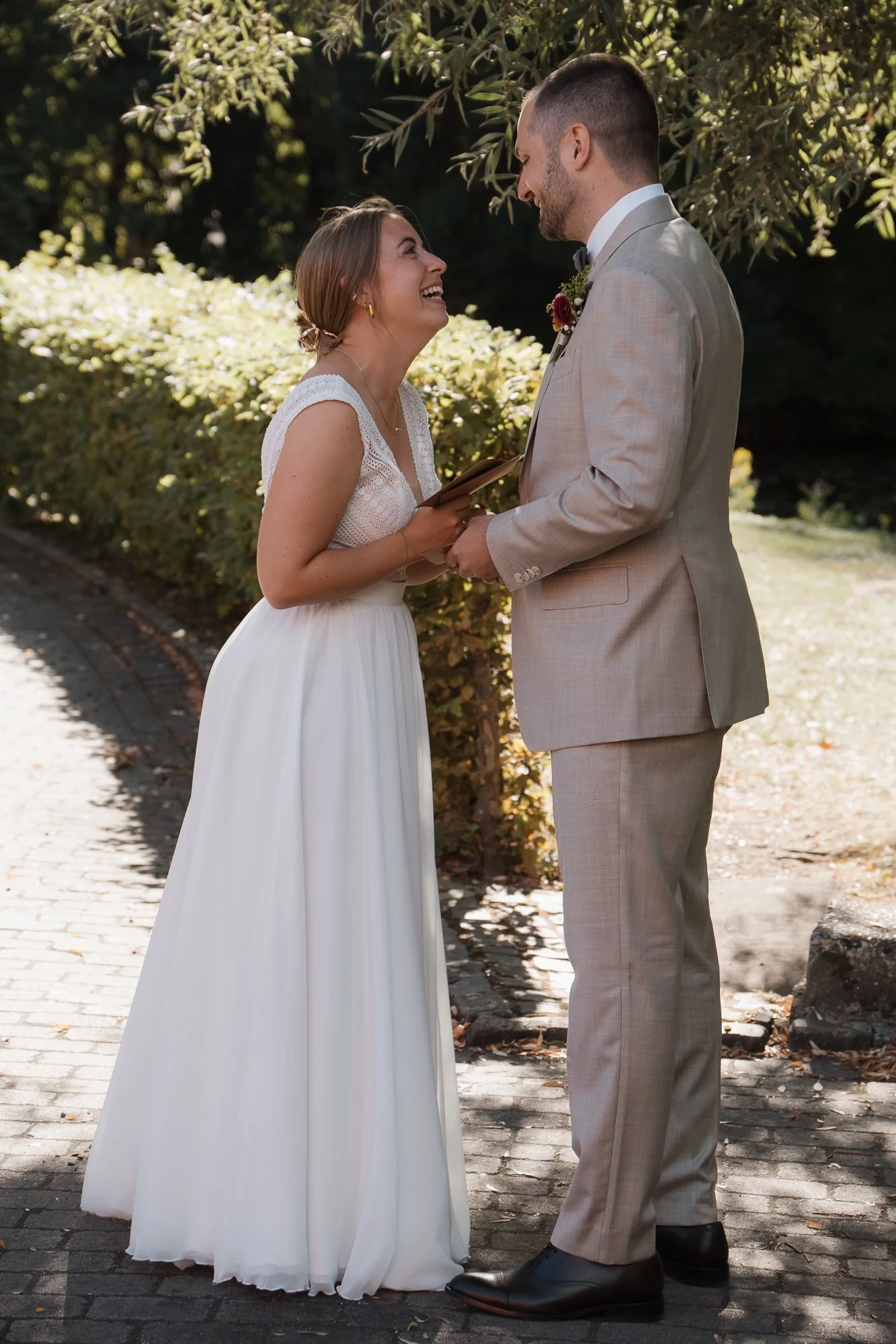 Ein Brautpaar steht sich bei einer standesamtlichen Trauung im Freien gegenüber, die Braut trägt ein weißes Brautkleid, der Bräutigam einen hellen Anzug, beide lachen und schauen sich liebevoll an.