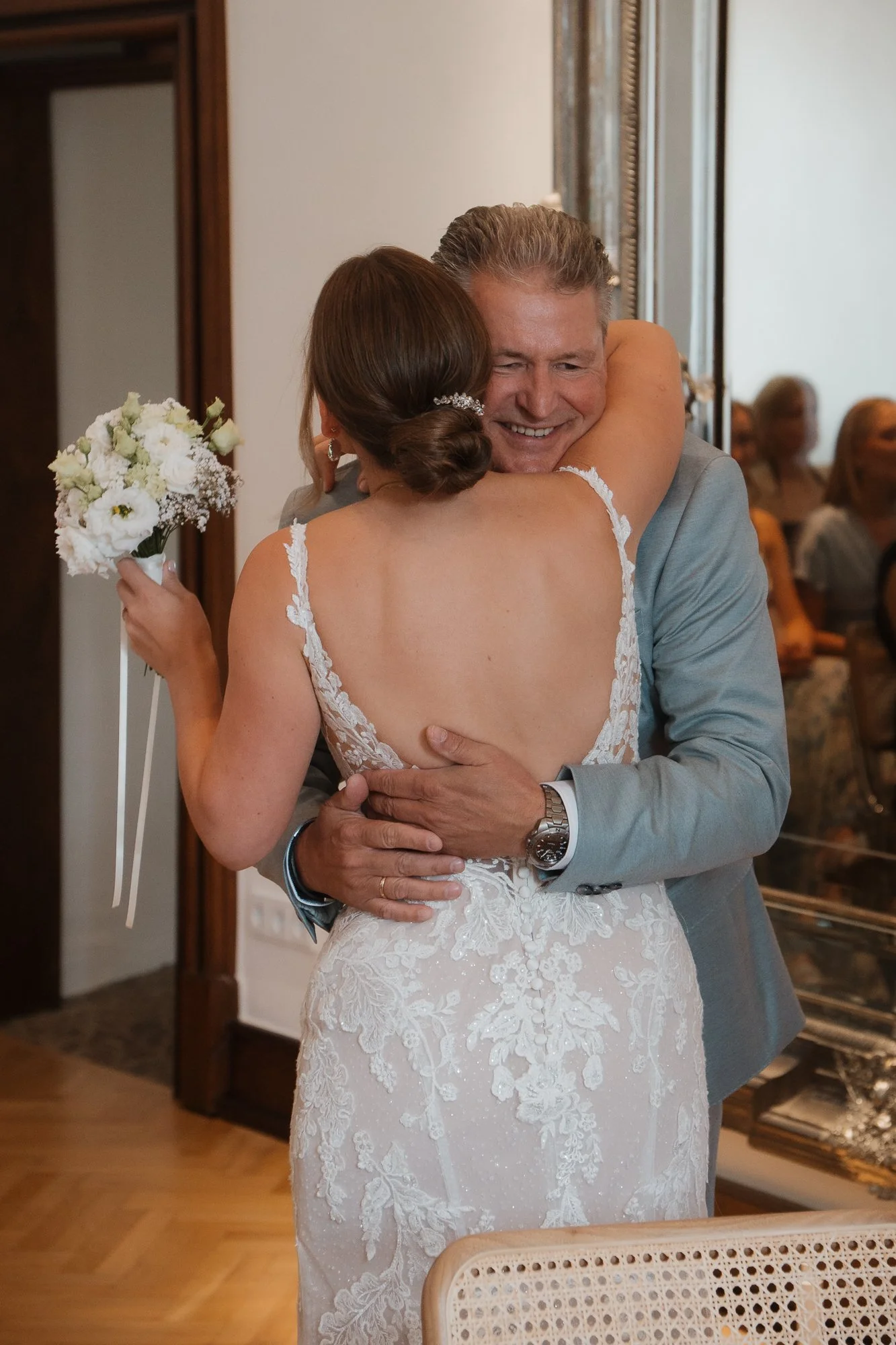 Ein Brautpaar bei einer Hochzeit, sie umarmen sich herzlich, die Braut trägt ein weißes Spitzenkleid und hält einen Blumenstrauß, im Hintergrund sind Gäste zu sehen.