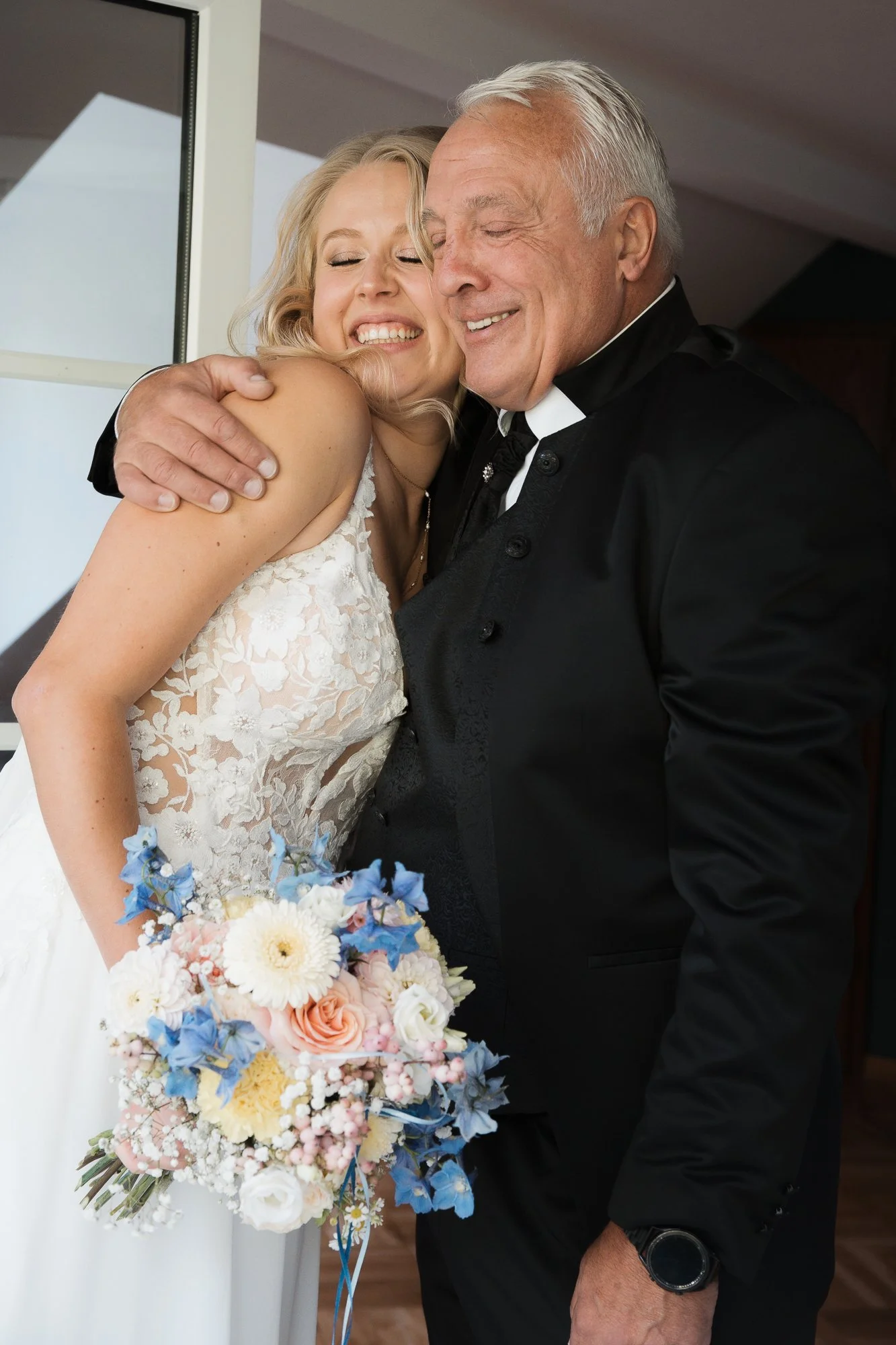 Ein junges Brautpaar umarmt sich und lächelt, während der Bräutigam einen älteren Mann in einem schwarzen Anzug umarmt, vermutlich an einem Hochzeitstag.