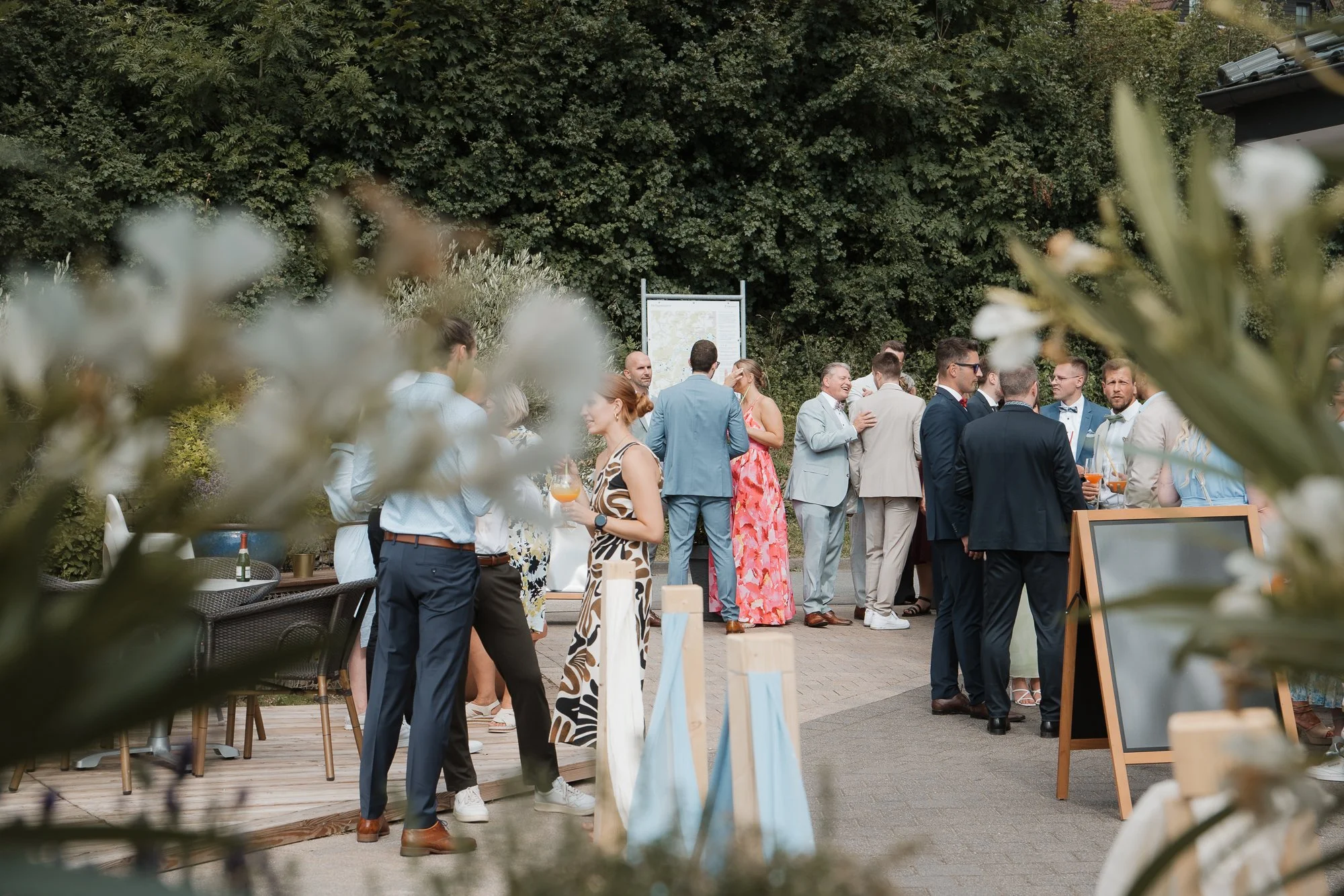 Menschen in formeller Kleidung bei einer Outdoor-Veranstaltung, die sich austauschen, mit einem Hintergrund aus Büschen und Bäumen, sowie einem steinernen Weg.