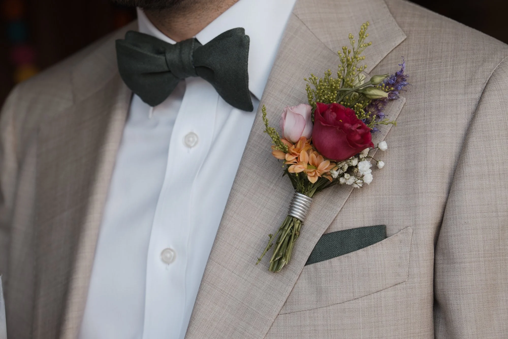 Nahaufnahme eines Hochzeitsanzugs mit Blumenboutonniere am Revers, bestehend aus roten, rosa und weißen Blumen, ergänzt durch grüne Zweige und kleine lila Blüten, getragen von einer Person in formeller Kleidung mit weißem Hemd und schwarzer Fliege.