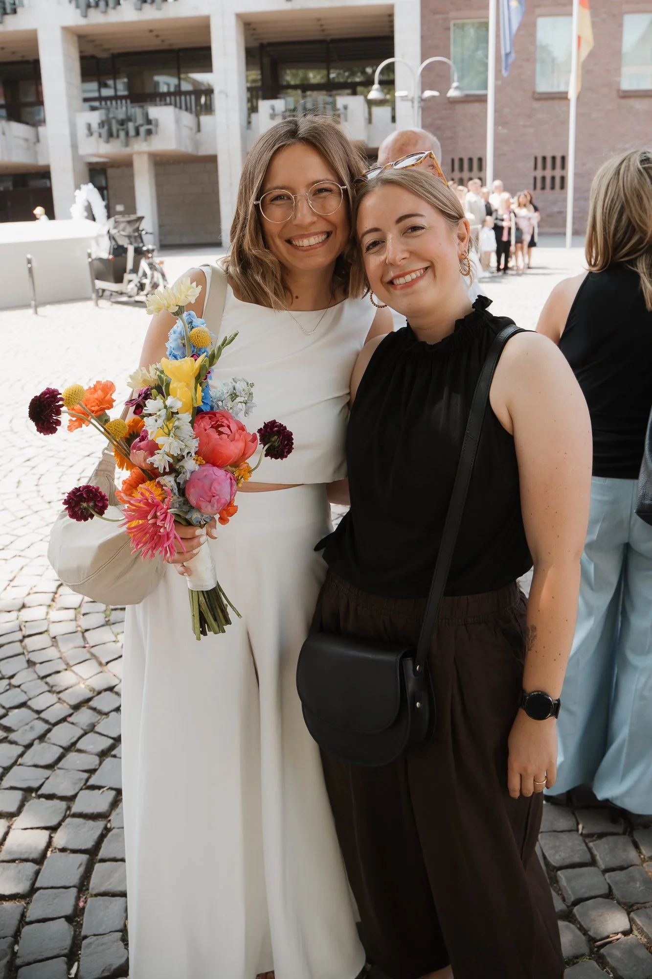 Zwei lachende Frauen, eine mit Brille in einem weißen Kleid, hält einen bunten Blumenstrauß, die andere in schwarzen Kleidung mit Tasche, stehen zusammen vor einem modernen Gebäude im Freien.