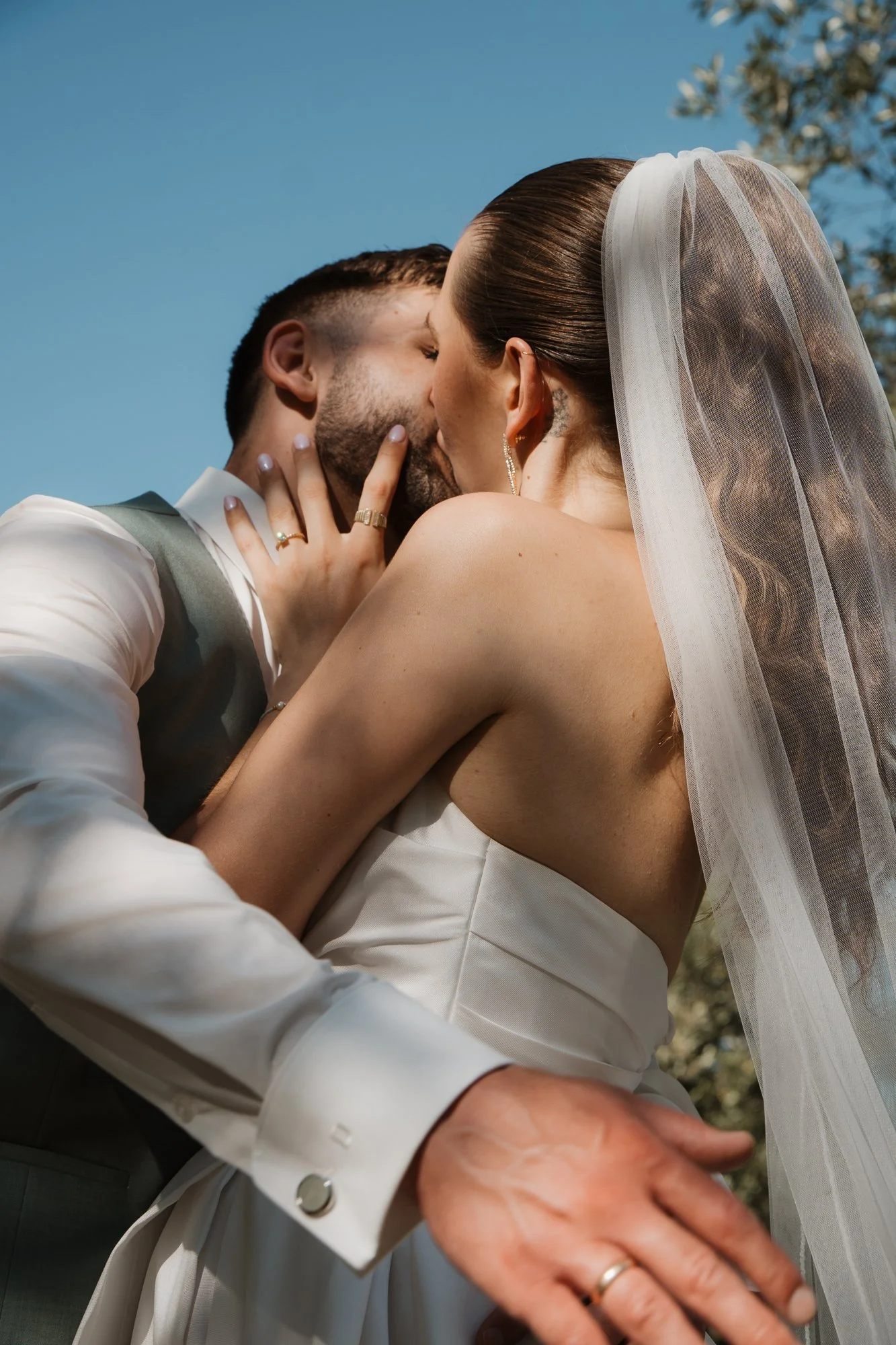Kussendes Paar bei einer Hochzeit, die Frau trägt ein Brautkleid und ein Schleier, der Mann trägt einen Anzug, im Hintergrund blauer Himmel und Blumen.