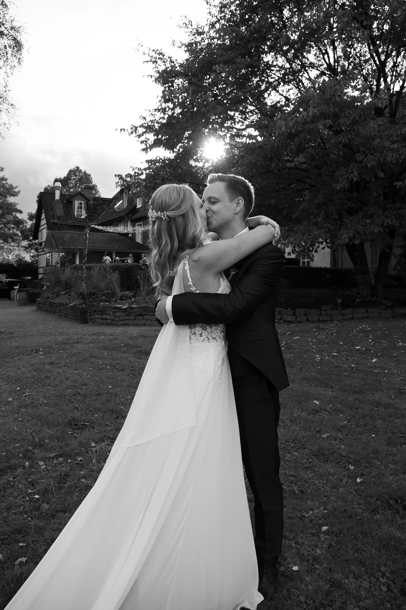 Braut und Bräutigam umarmen sich und küssen sich bei Sonnenuntergang im Garten, mit Bäumen und einem Haus im Hintergrund.