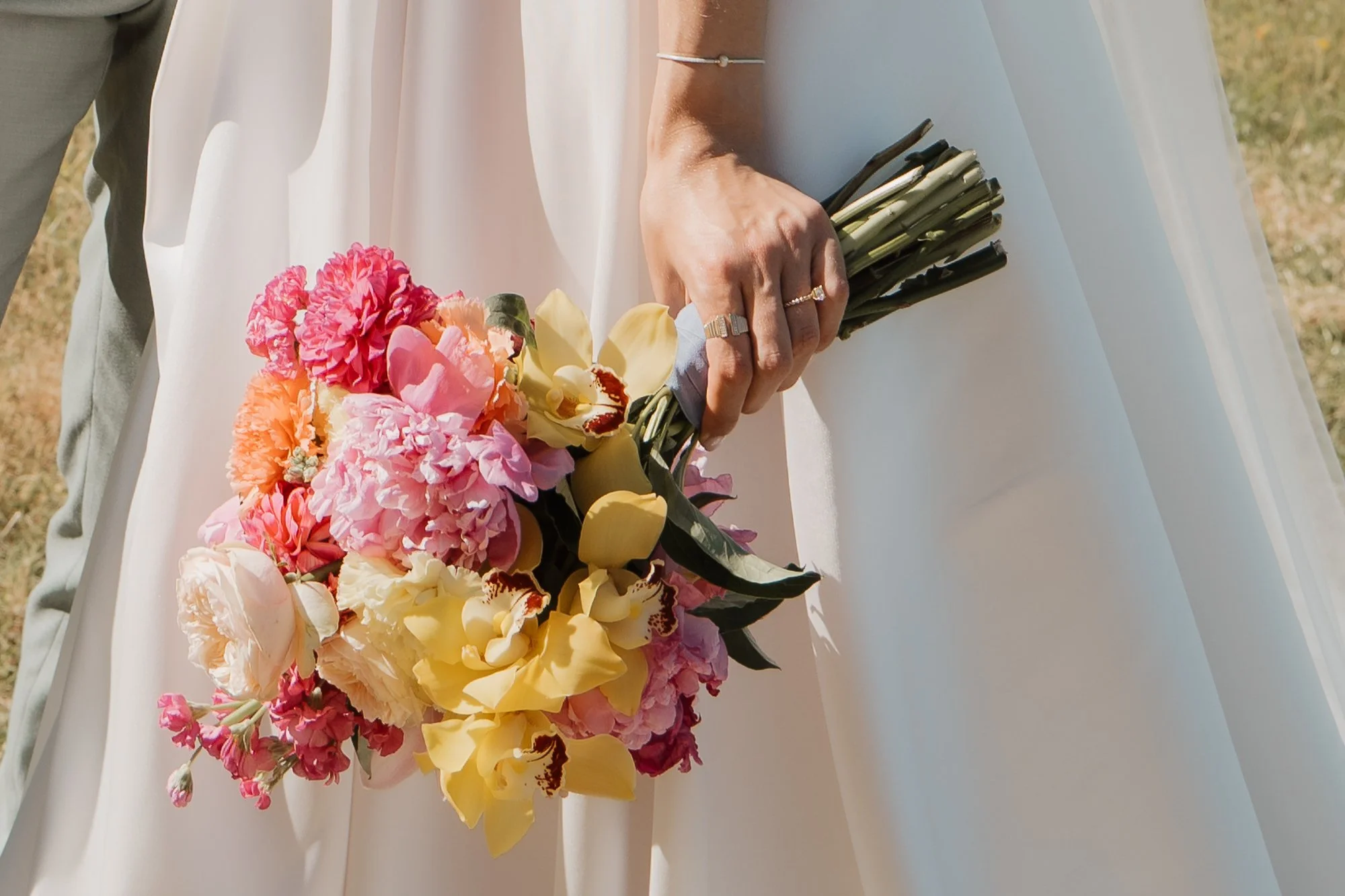 Nahaufnahme einer Hand, die einen Brautstrauß mit bunten Blumen hält, während eine Braut in einem weißen Kleid steht.