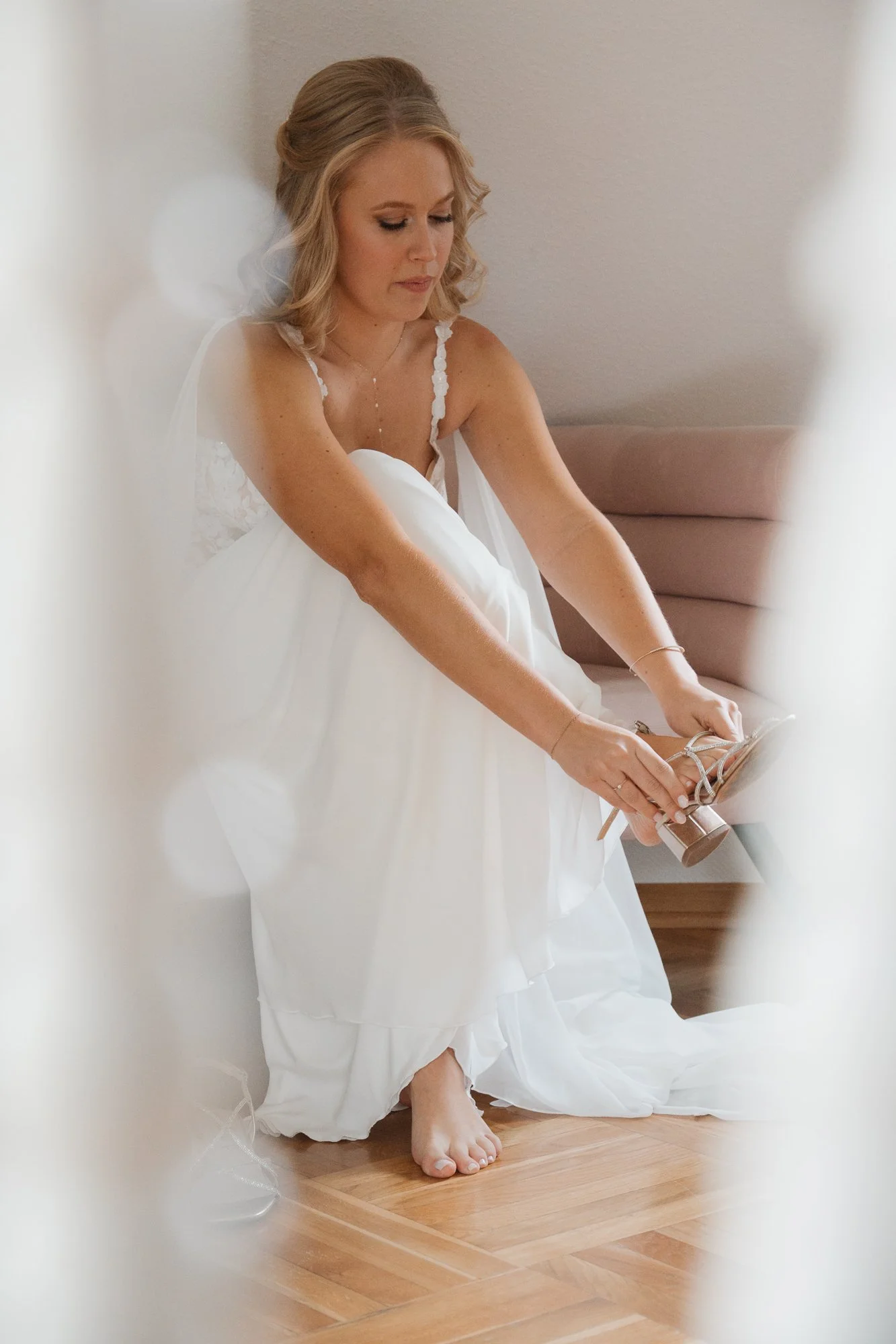 Braut sitzt auf einem Stuhl und zieht ihre weißen Hochzeitsschuhe an, im Hintergrund eine rote Couch, der Raum hat eine helle Wand.