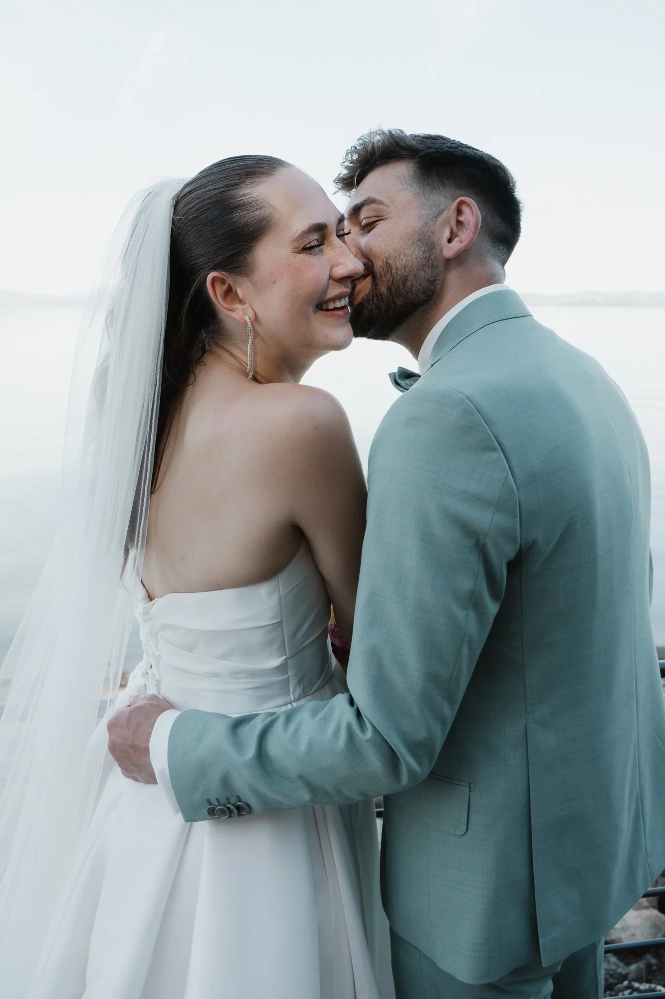 Ein Brautpaar in Hochzeitskleidung, das sich an einem Wasser im Freien küsst.