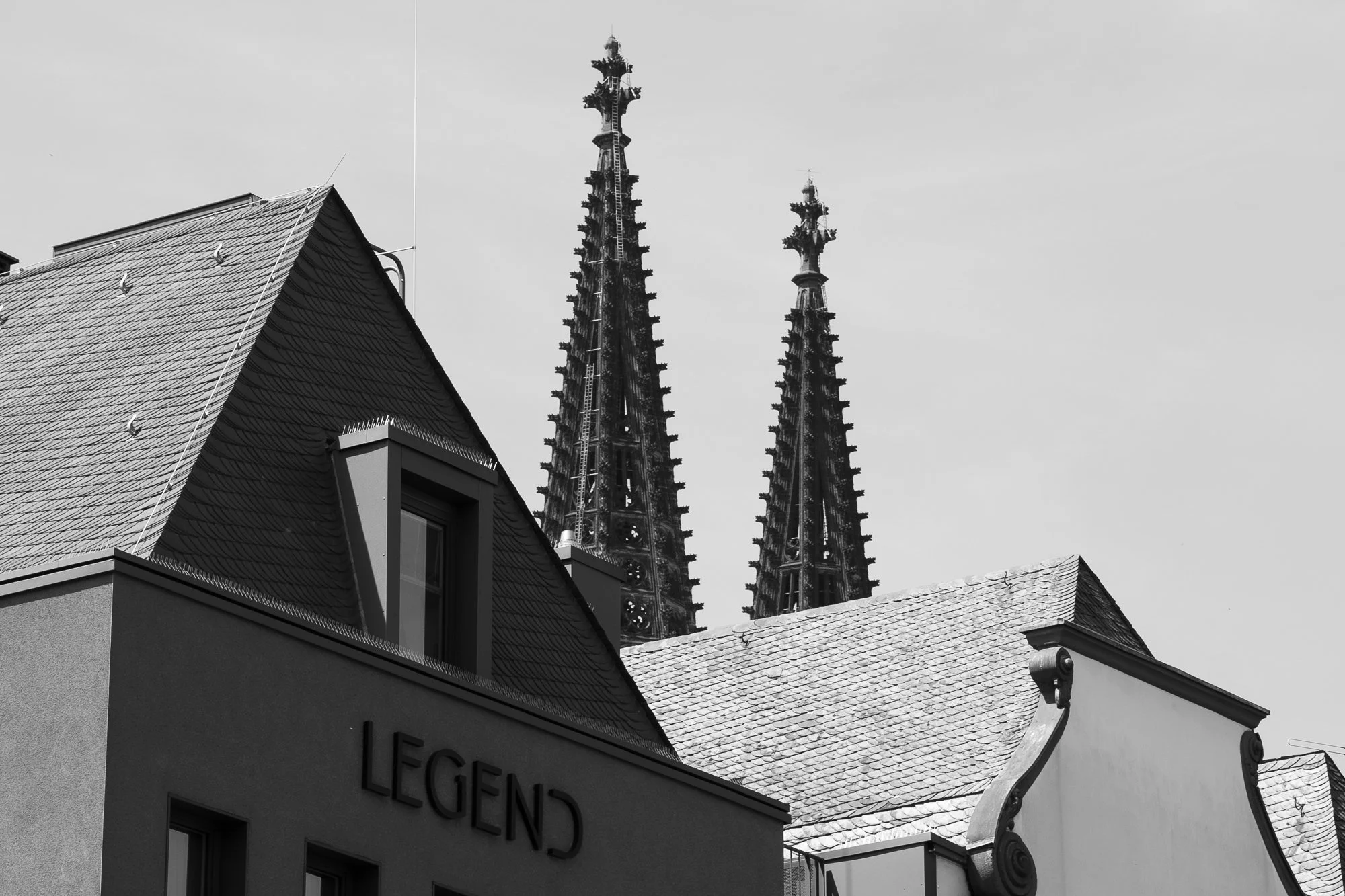 Schwarz-Weiß-Bild zeigt die Dächer eines modernen Gebäudes mit dem Schriftzug 'LEGEND' im Vordergrund und die spitzen Türme einer gotischen Kathedrale im Hintergrund.