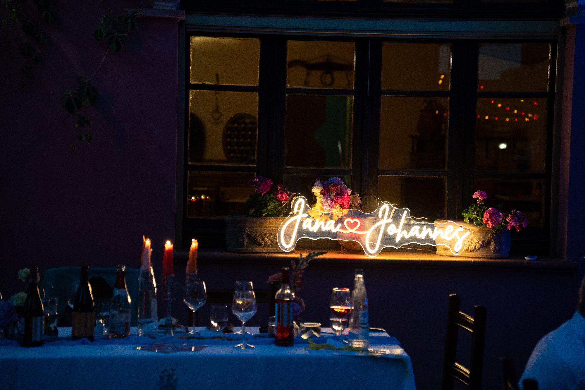Ein festlich gedeckter Tisch mit mehreren Weingläsern und Kerzen vor einem Fenster. Auf dem Fensterbrett leuchtet ein Neon-Schild mit der Aufschrift 'Jana ❤️ Johannes' und kleinen Blumen.