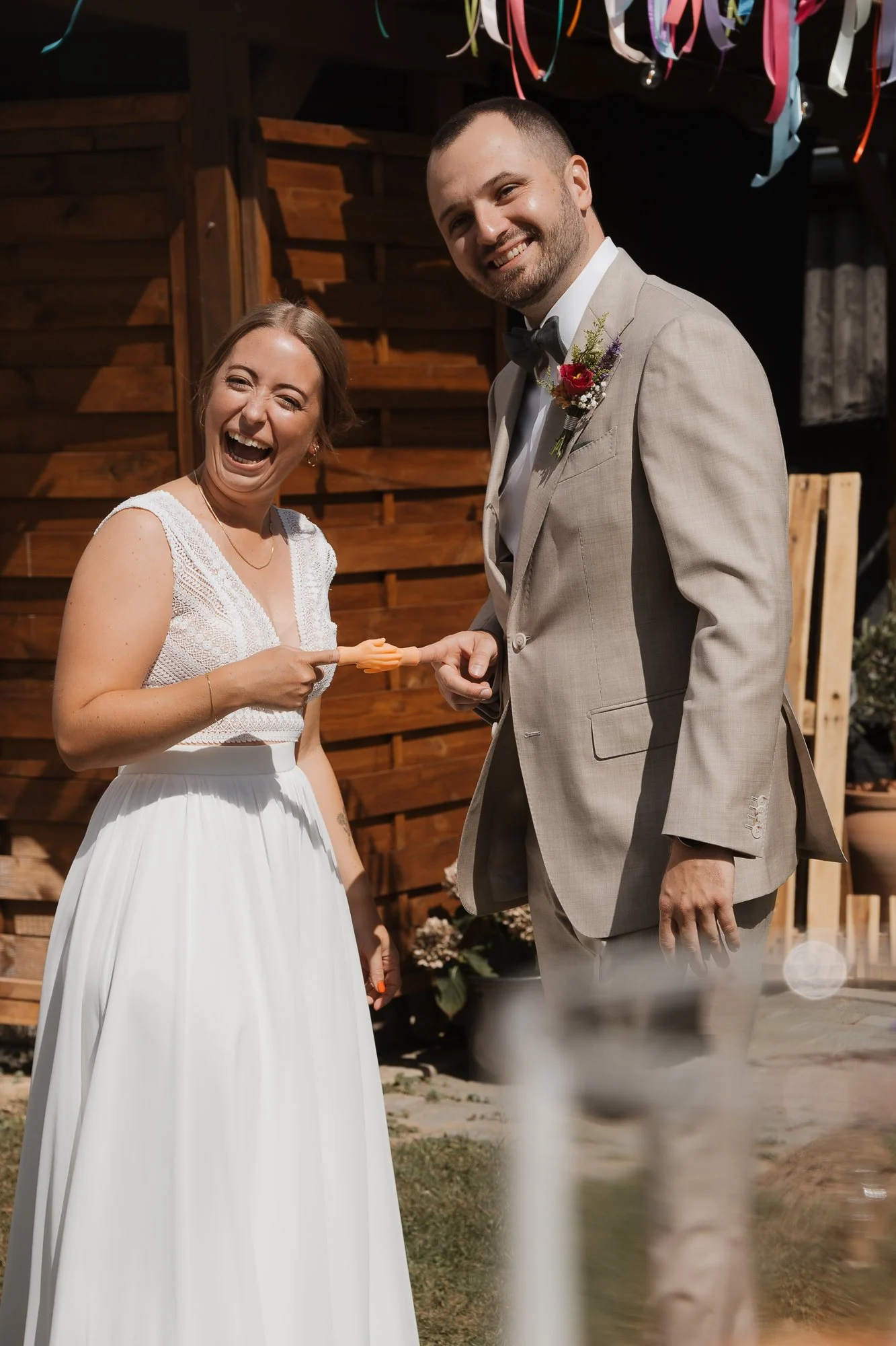 Ein lachendes Hochzeitspaar bei ihrer Zeremonie im Freien, die Frau hält einen Orangenschwamm, im Hintergrund eine Holzwand und bunte Bänder hängen runter.