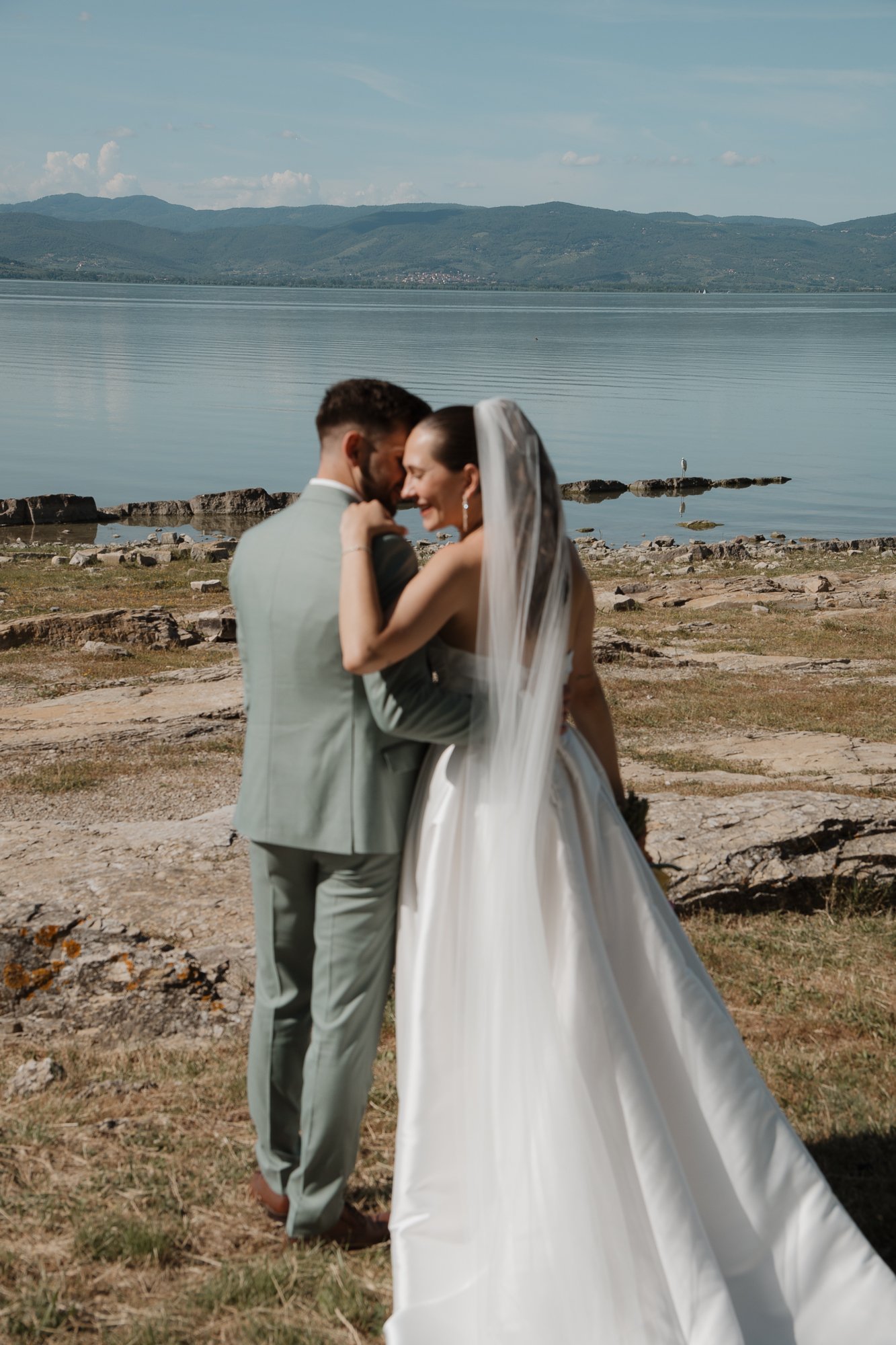 Ein Paar in Hochzeitskleidung steht am Ufer eines Sees, umgeben von Bergen. Sie berühren sich zärtlich und lachen, während die Braut einen Schleier trägt.
