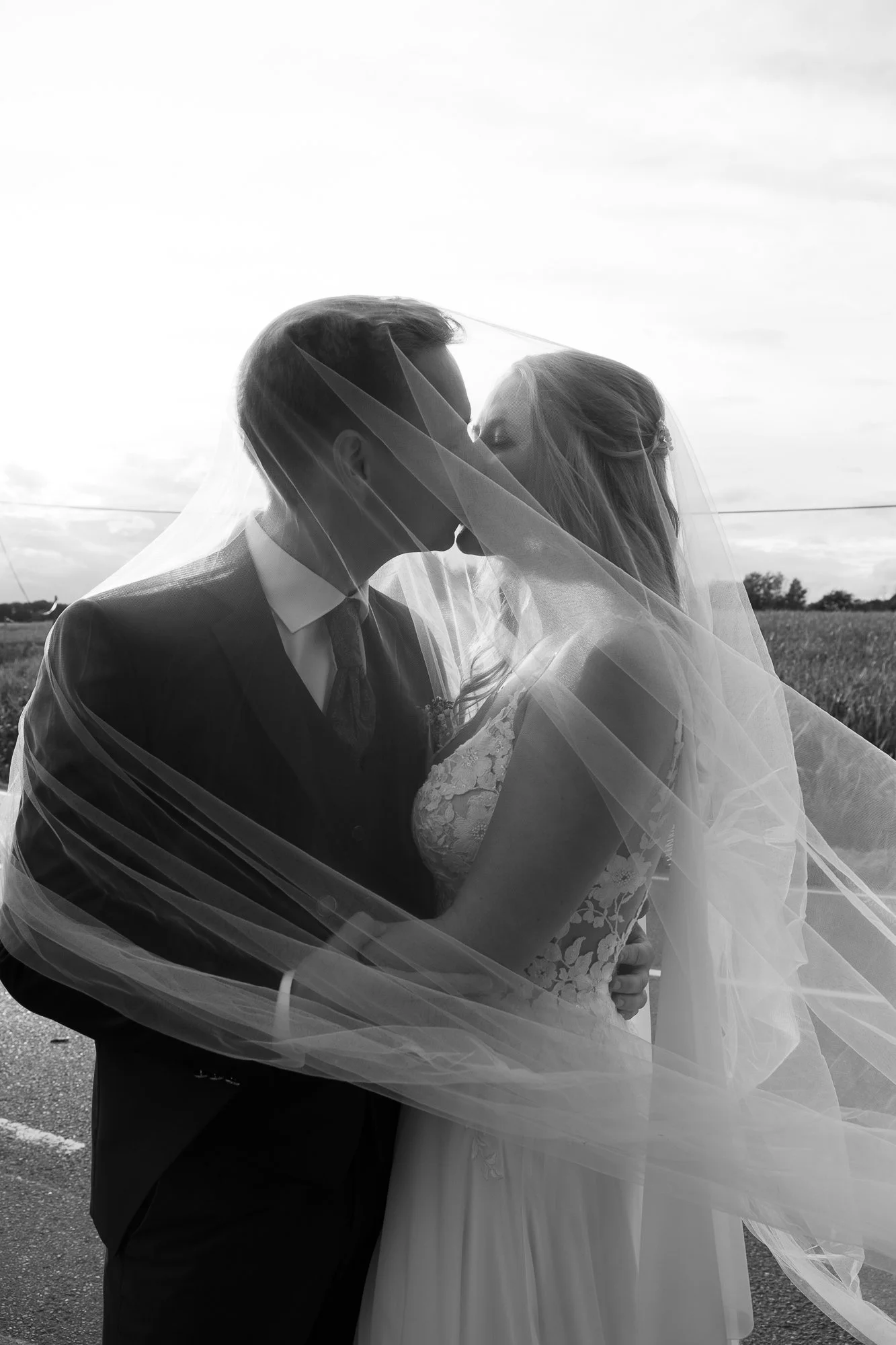 Ein Brautpaar küsst sich unter einem Schleier im Freien bei Sonnenuntergang, schwarz-weiß Aufnahme.