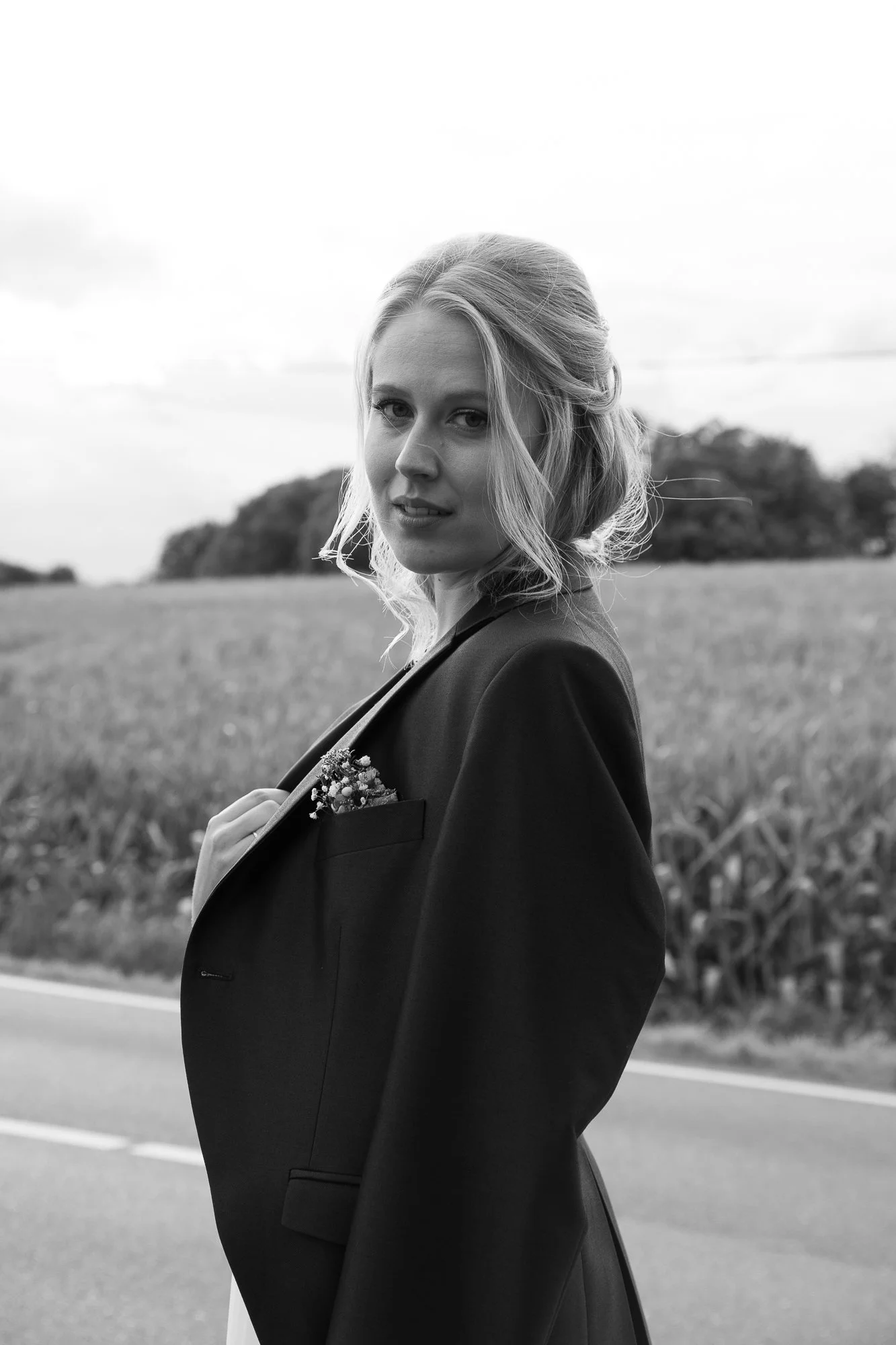 Schwarz-Weiß-Foto einer Frau in einem Anzug mit einer kleinen Blume in der Brusttasche, im Freien an einer Landstraße mit einem Feld im Hintergrund.