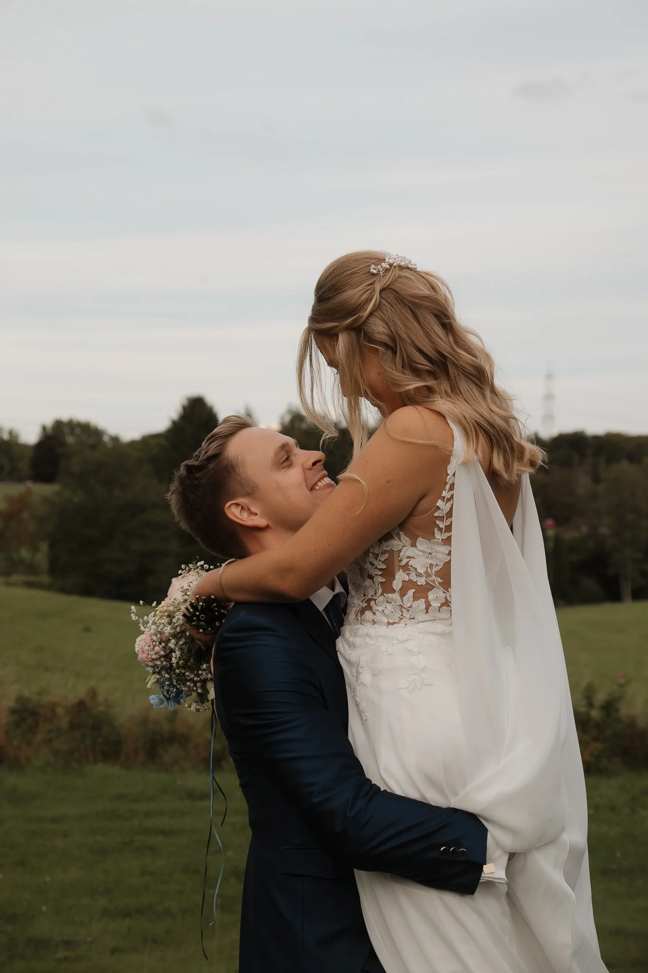 Ein Brautpaar bei der Hochzeitszeremonie im Freien, der Bräutigam hebt die braut hoch, beide lächeln glücklich, im Hintergrund ein grüner Garten unter einem bewölkten Himmel.