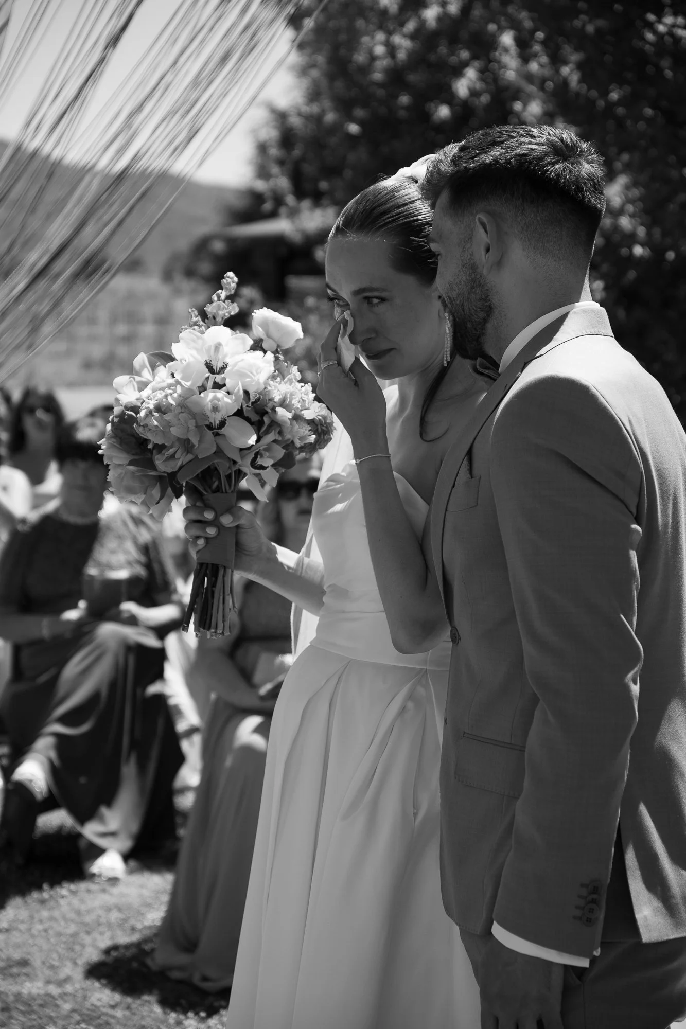 Ein Brautpaar während einer Hochzeit, das scheinbar emotional berührt ist, mit Gästen im Hintergrund, im Freien bei Sonnenschein.