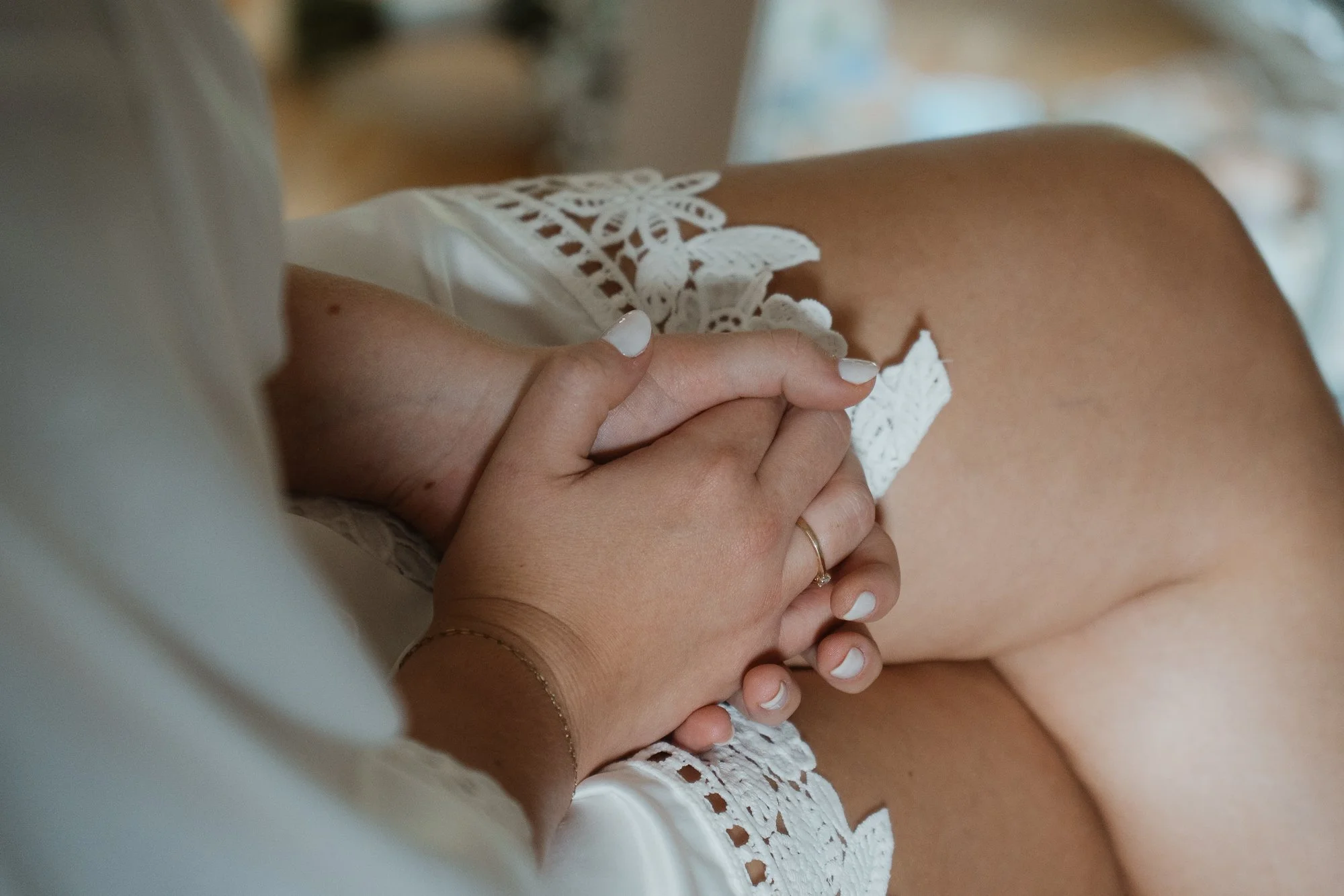 Nahaufnahme von Händen, die auf einem weißen Spitzenstoff ruhen, mit einem Ring am Finger, in einem intimen Moment.