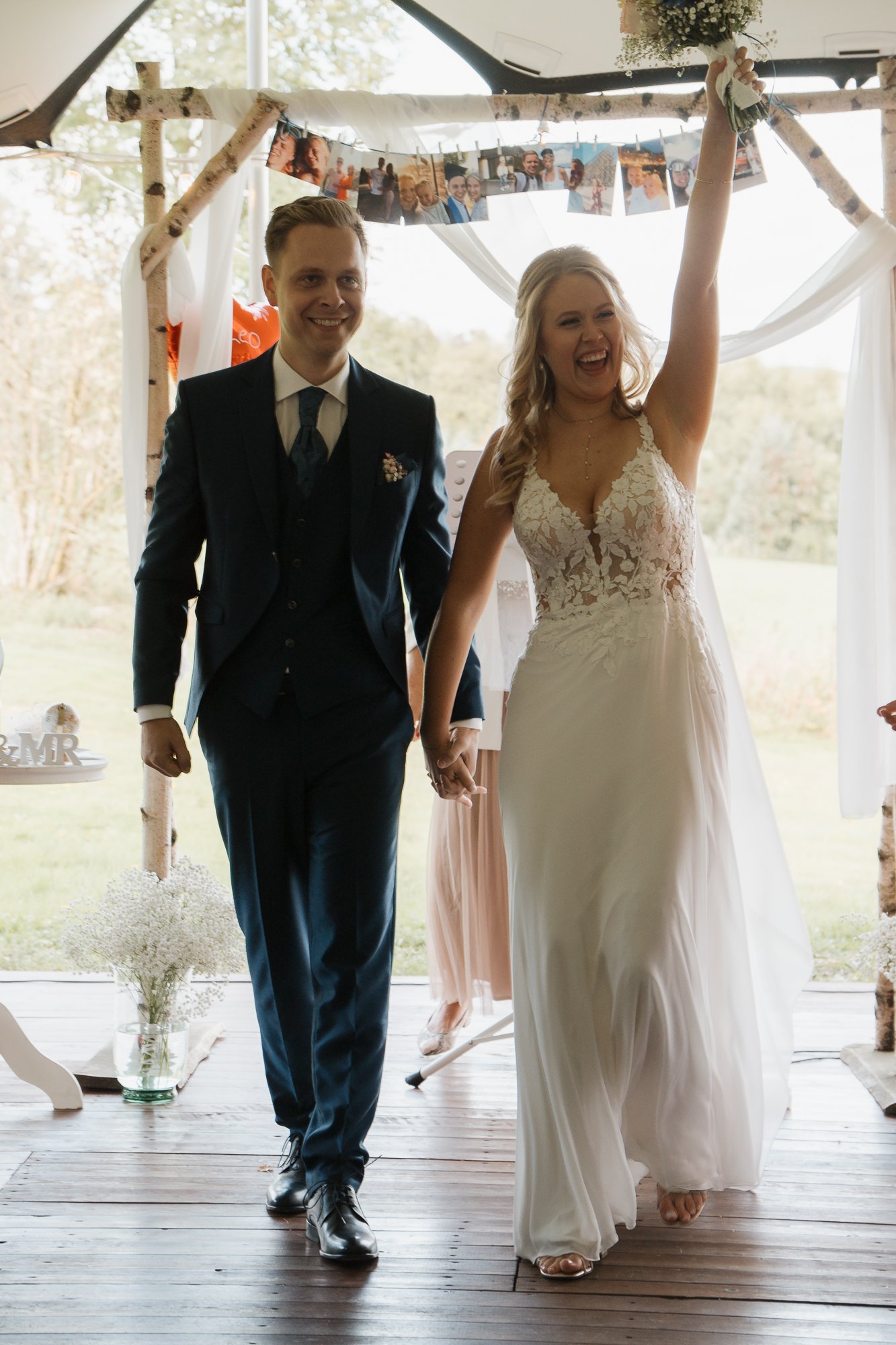 Braut und Bräutigam beim Heiratsauftakt, Hand in Hand, lachend, in einer festlichen Umgebung mit Blumen und Fotos im Hintergrund.