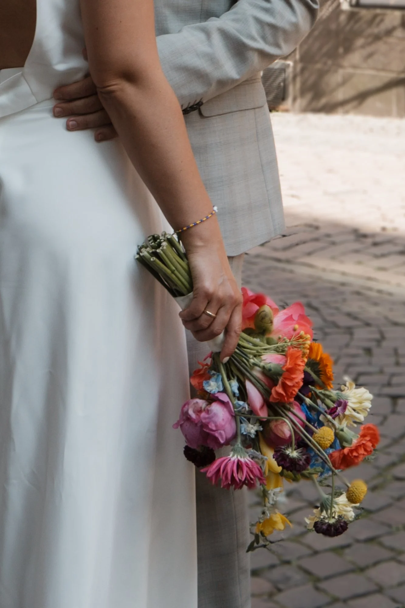 Ein Hochzeitsstrauß aus bunten Blumen, gehalten von einer Person in einem weißen Kleid, während jemand anderes eine graue Anzughose trägt.