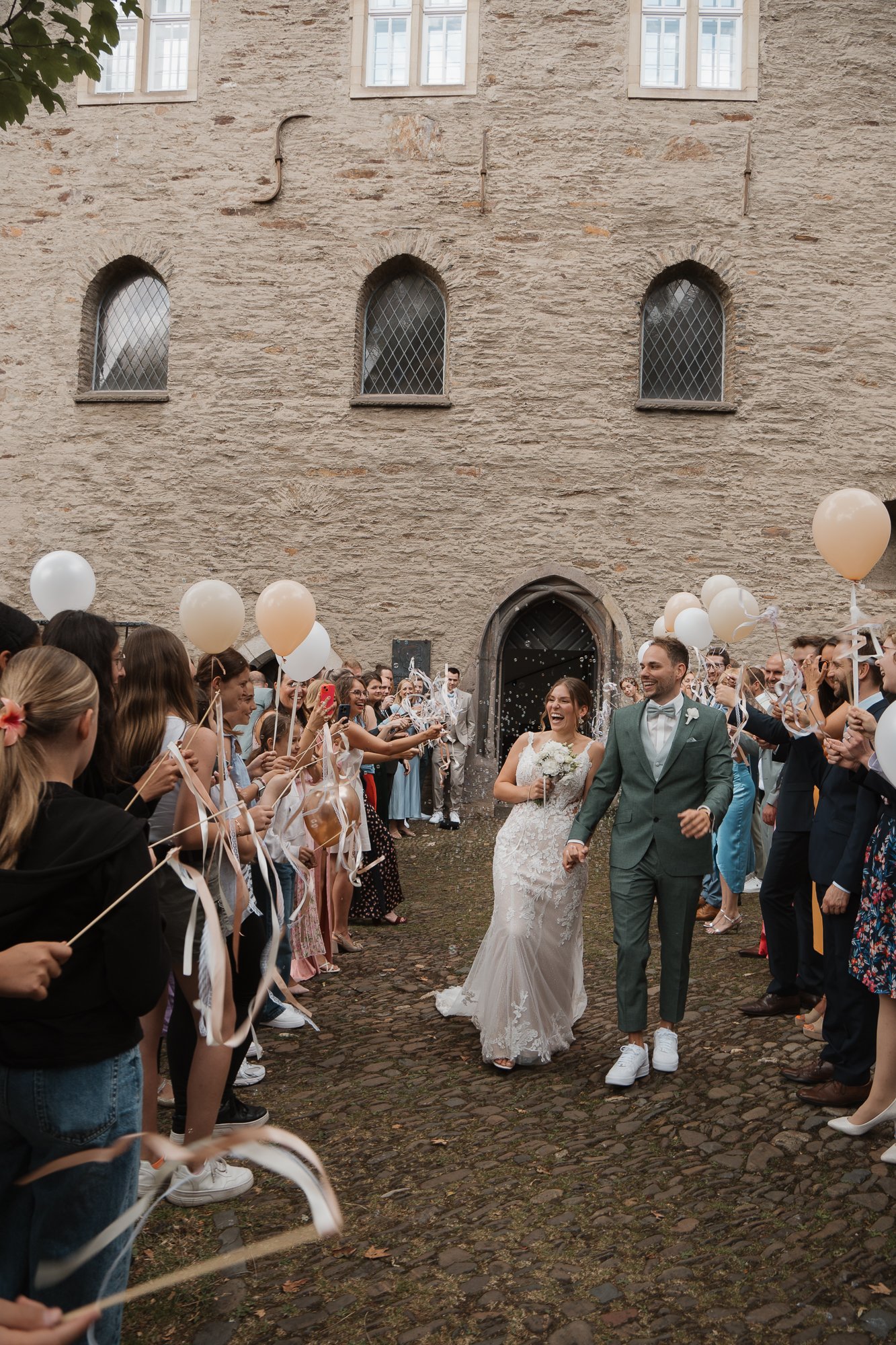 Ein Brautpaar läuft Hand in Hand durch eine Menschenmenge bei einer Hochzeit vor einer alten Steinmauer. Die Gäste werfen gemeinsam Konfetti und Ballons in die Luft, alle sind fröhlich und feiern den Moment.