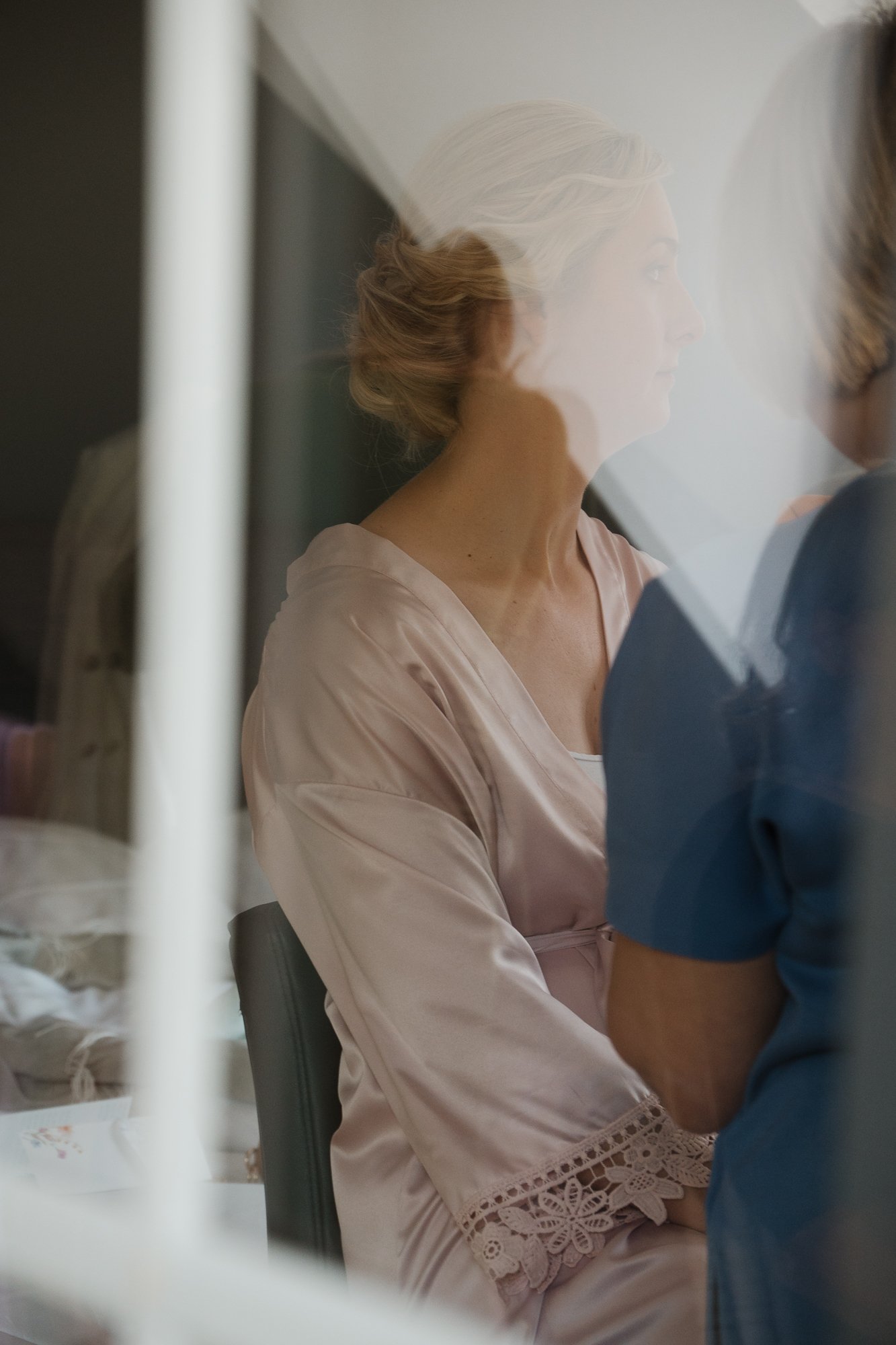 Eine Frau mit blonden Haaren, die in eine Schulter geklammert sind, sitzt durch ein Fenster betrachtet. Es ist ein Spiegel oder eine Glasscheibe zwischen ihr und dem Betrachter.