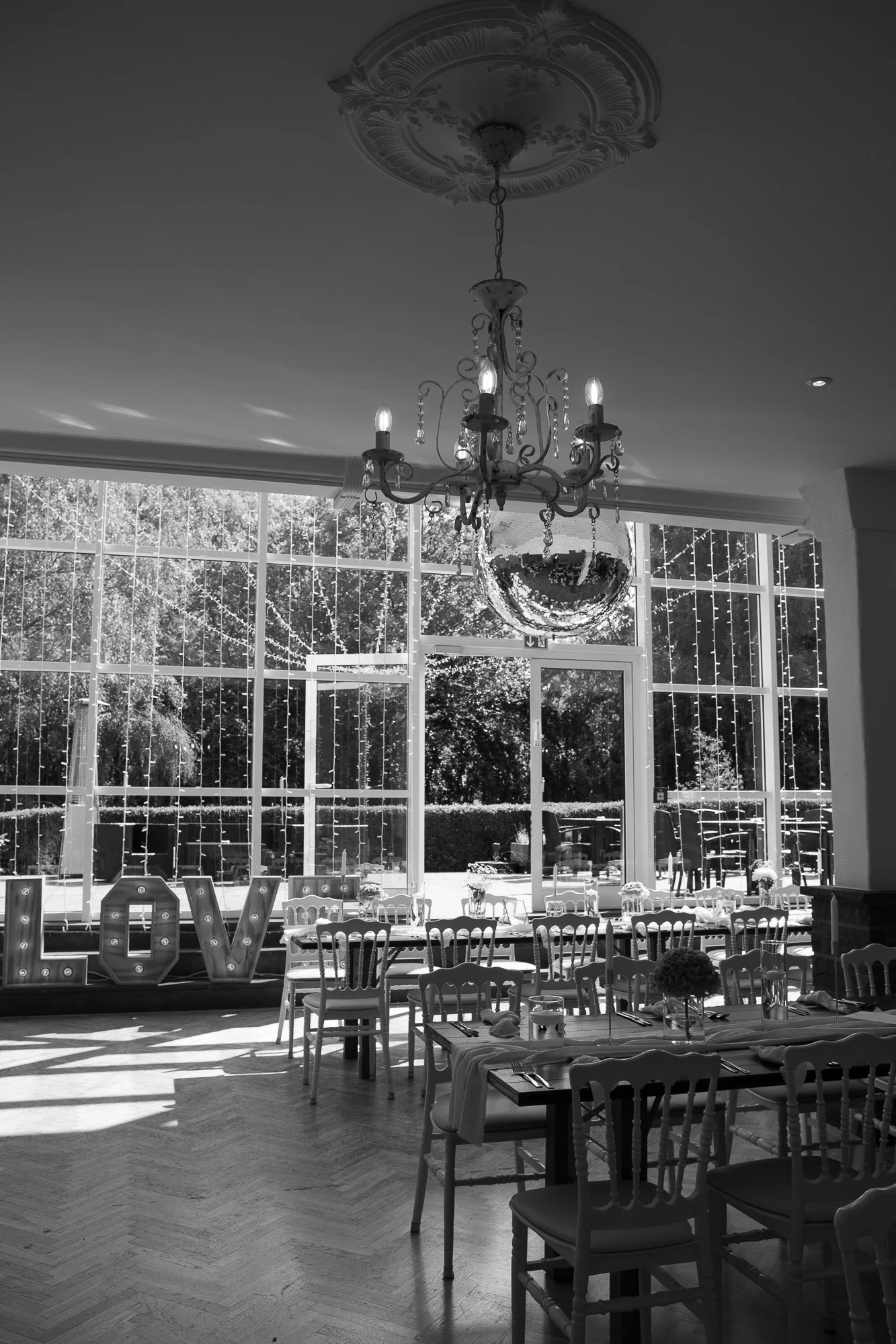 Innenansicht eines festlich gedeckten Raums mit großem Fenster, einer Kristallkugel und einem Kronleuchter, mit Blumenschmuck auf den Tischen.