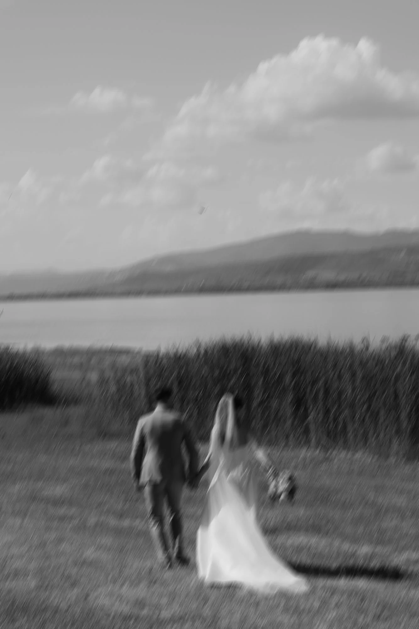 Ein Paar in formeller Kleidung, vermutlich Braut und Bräutigam, läuft gemeinsam über eine Wiese mit einem Hund, im Hintergrund ein See und Berge, die Szene ist unscharf und in Schwarz-Weiß.