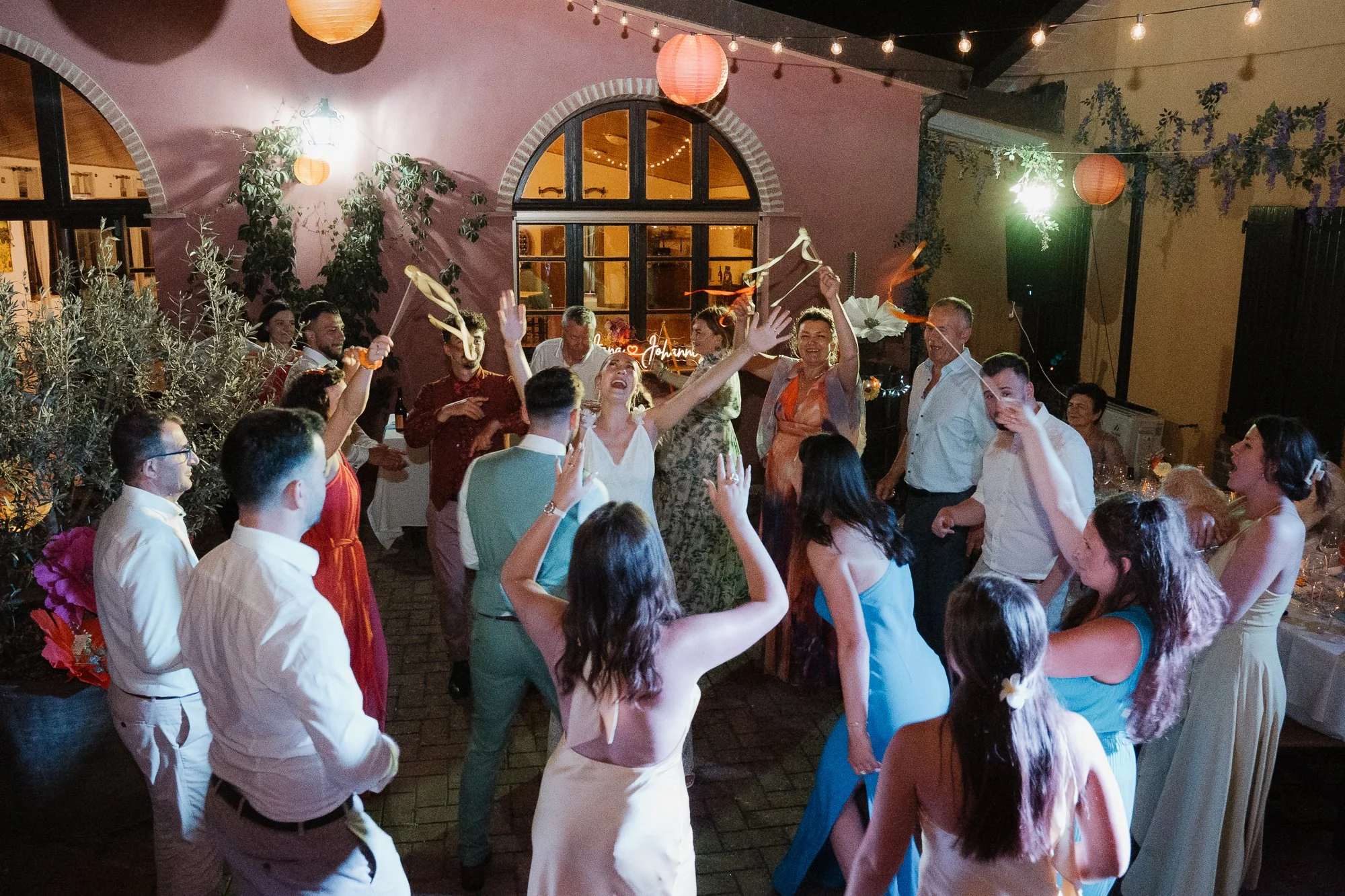 Menschen feiern mit Tanz und Gesang bei einer Party im Garten, umgeben von Lichterketten und bunten Lampions.