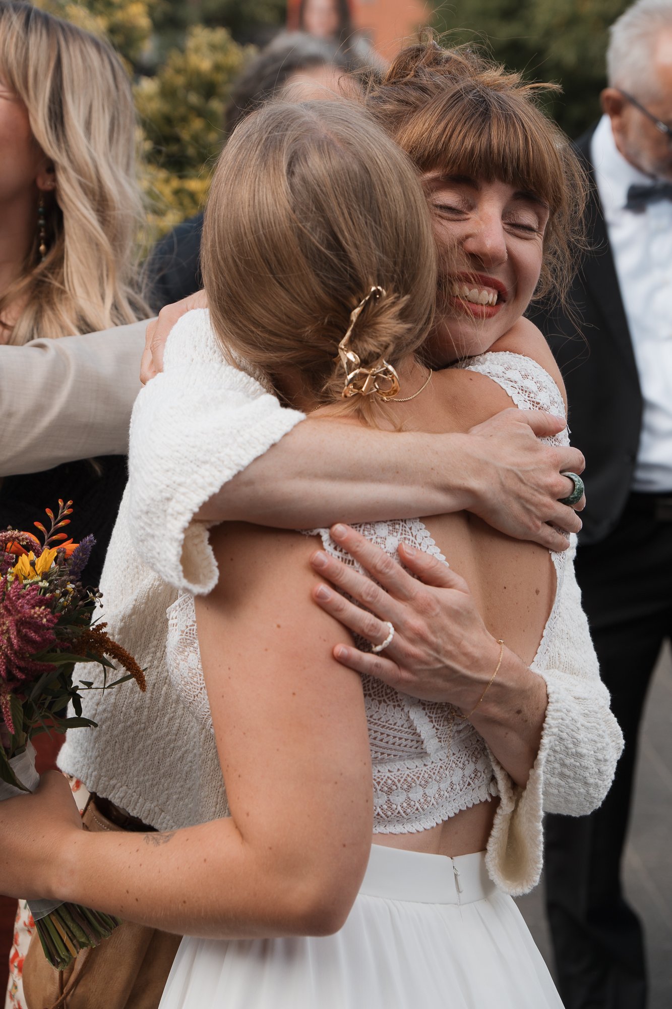 Zwei Frauen umarmen sich glücklich bei einer Feier, im Hintergrund sind weitere Menschen und Natur zu sehen.