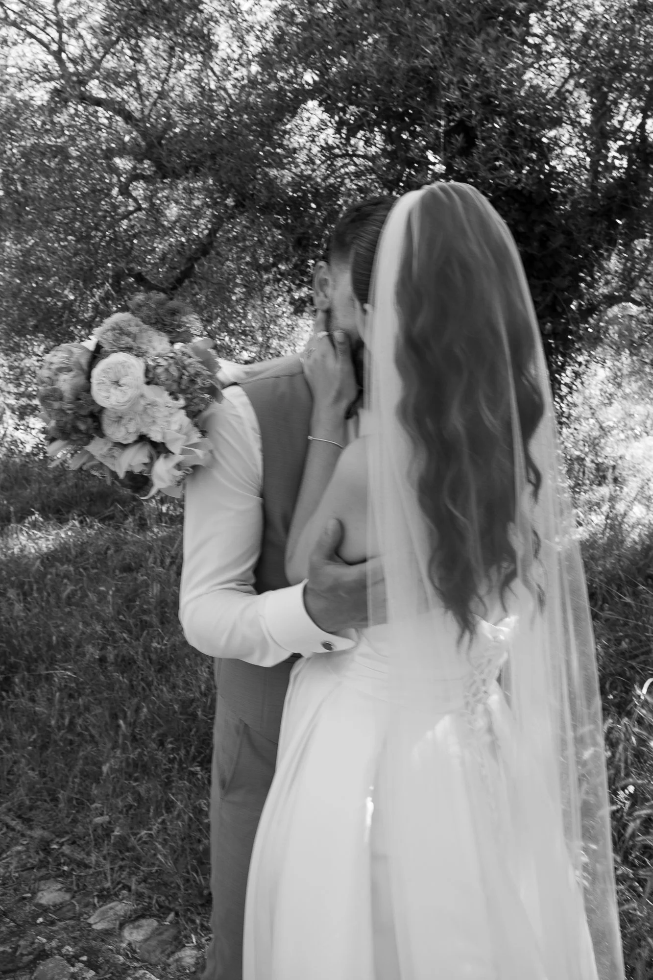 Ein Brautpaar teilt einen Kuss im Freien, die Braut trägt ein weißes Kleid und einen Schleier, der Bräutigam trägt ein Hemd und Weste, im Hintergrund Bäume.