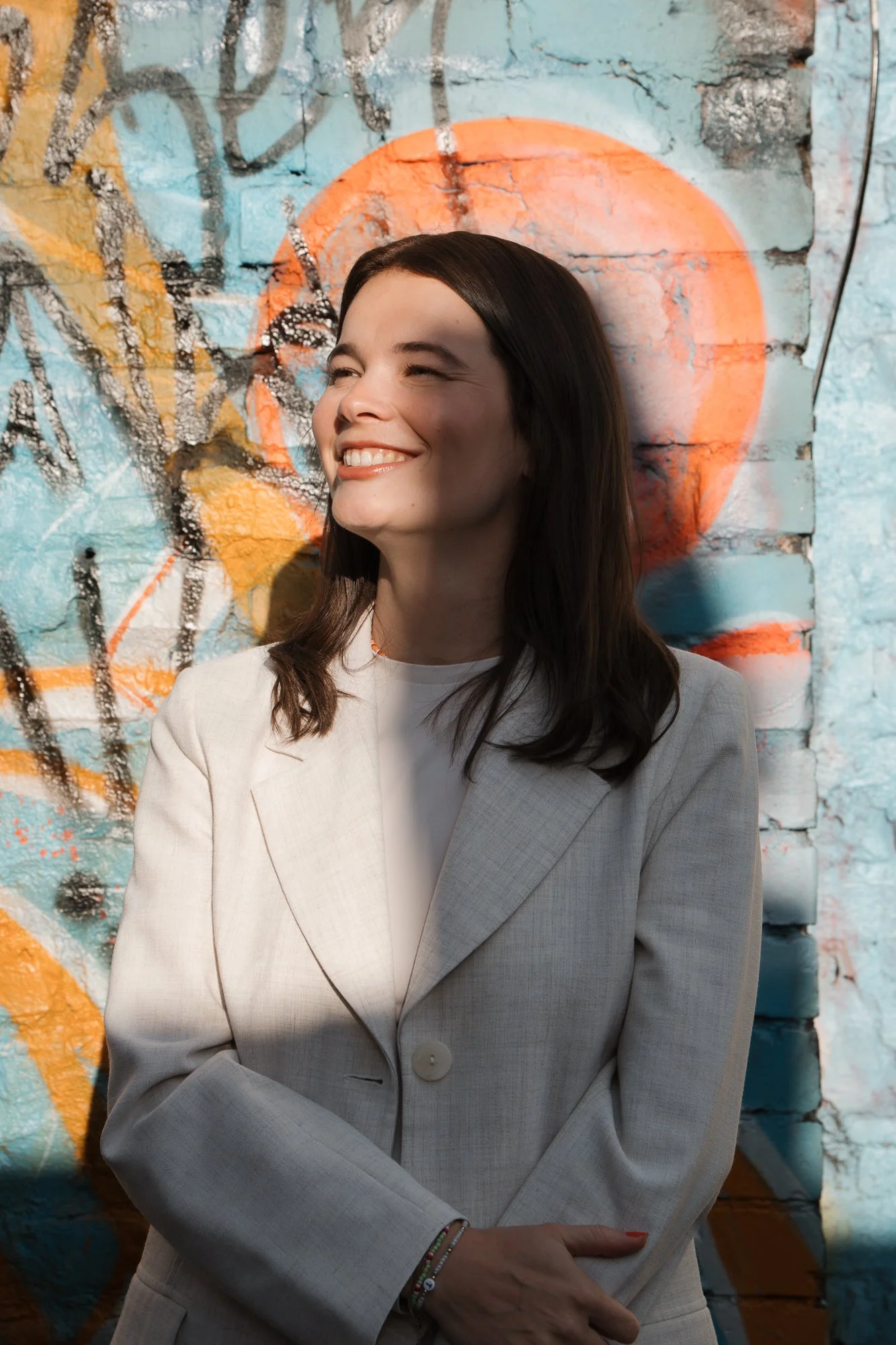 Eine lachende Frau mit dunklem Haar in einem weißen Blazer vor einer bunten Graffiti-Wand.