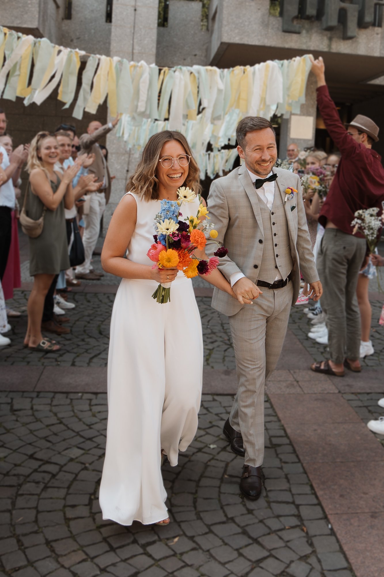 Hochzeitspaar, Hand in Hand, bei einer festlichen Feier im Freien, mit Gästen, die Blumen und Papierstreifen unterstützen.