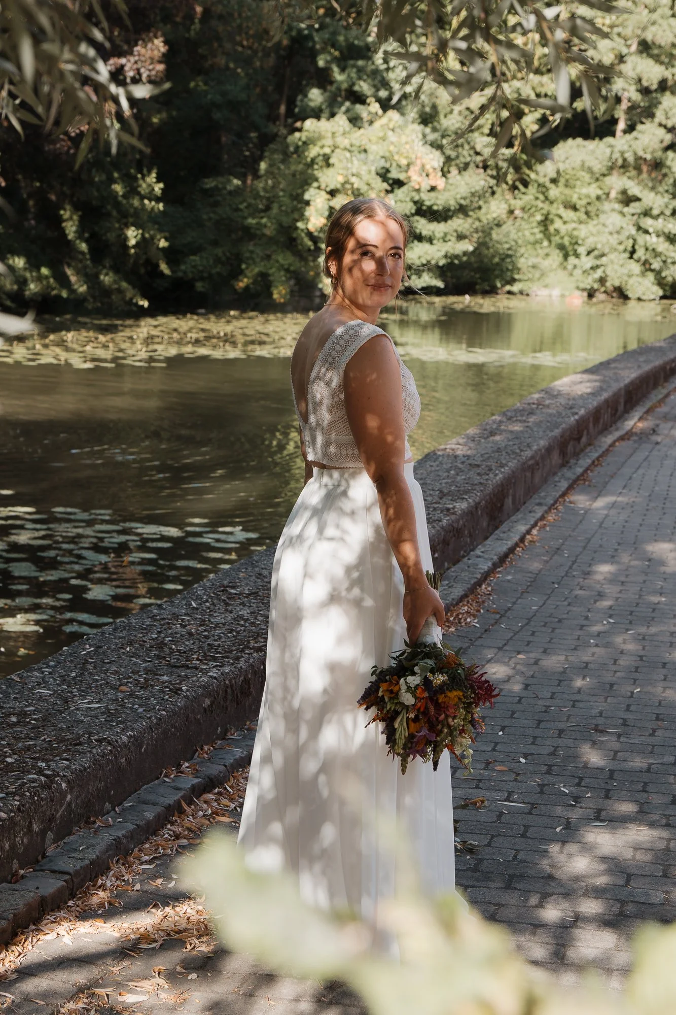 Junge Frau in Hochzeitskleid mit Blumenstrauß am Wasser in einem Park.