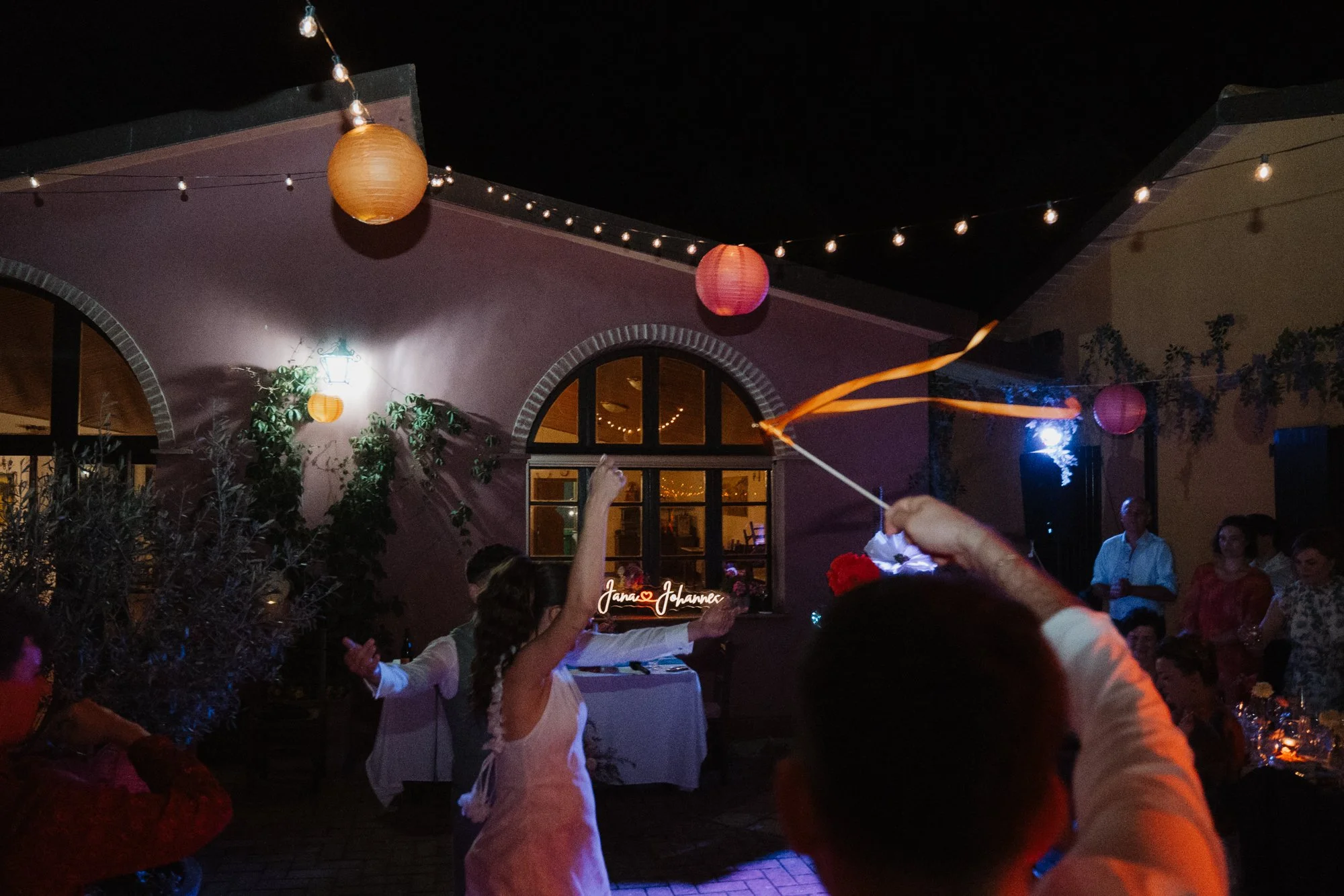 Menschen feiern bei Nacht im Außenbereich einer Location, mit bunten Laternen und Lichterketten. Es tanzt eine Frau in einem weißen Kleid, während ein Gast einen Luftballon hält.