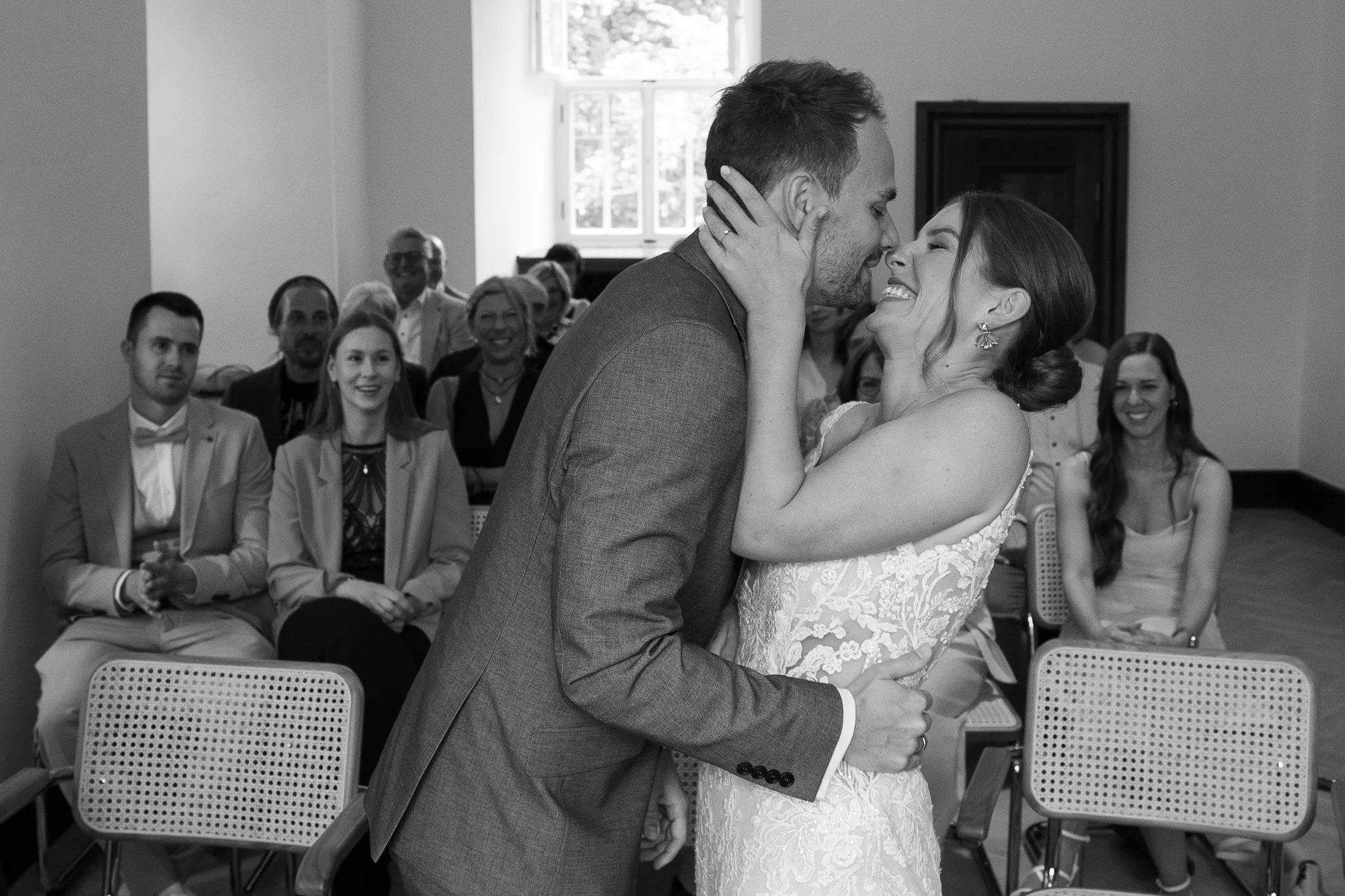 Ein glückliches Brautpaar küsst sich bei ihrer Hochzeit, umgeben von Freunden und Familie im Saal, schwarz-weiß Aufnahme.