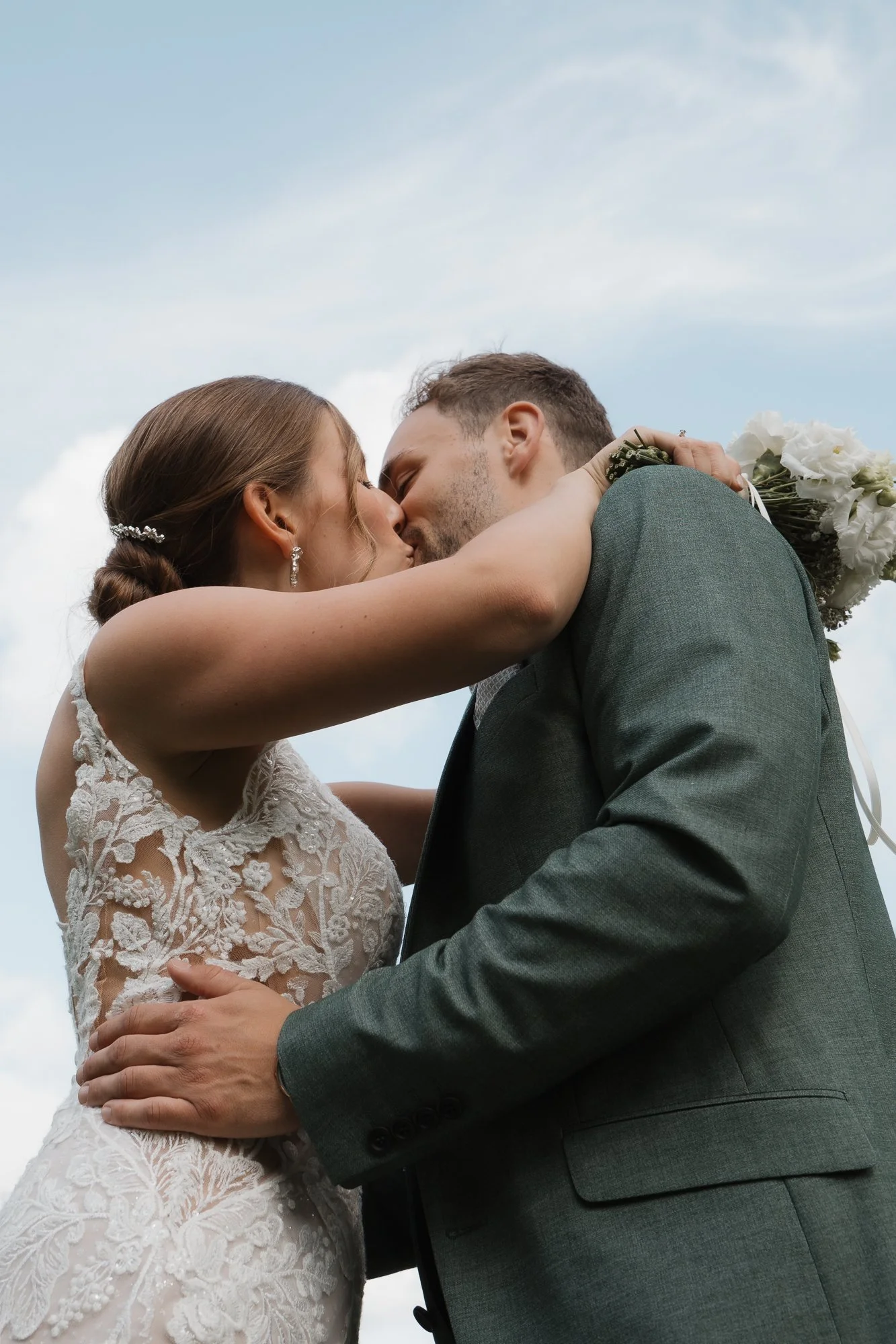 Ein Brautpaar küsst sich bei einem Hochzeitsfoto im Freien, die Braut trägt ein weißes Spitzenkleid und das Paar hält einen Blumenstrauß.