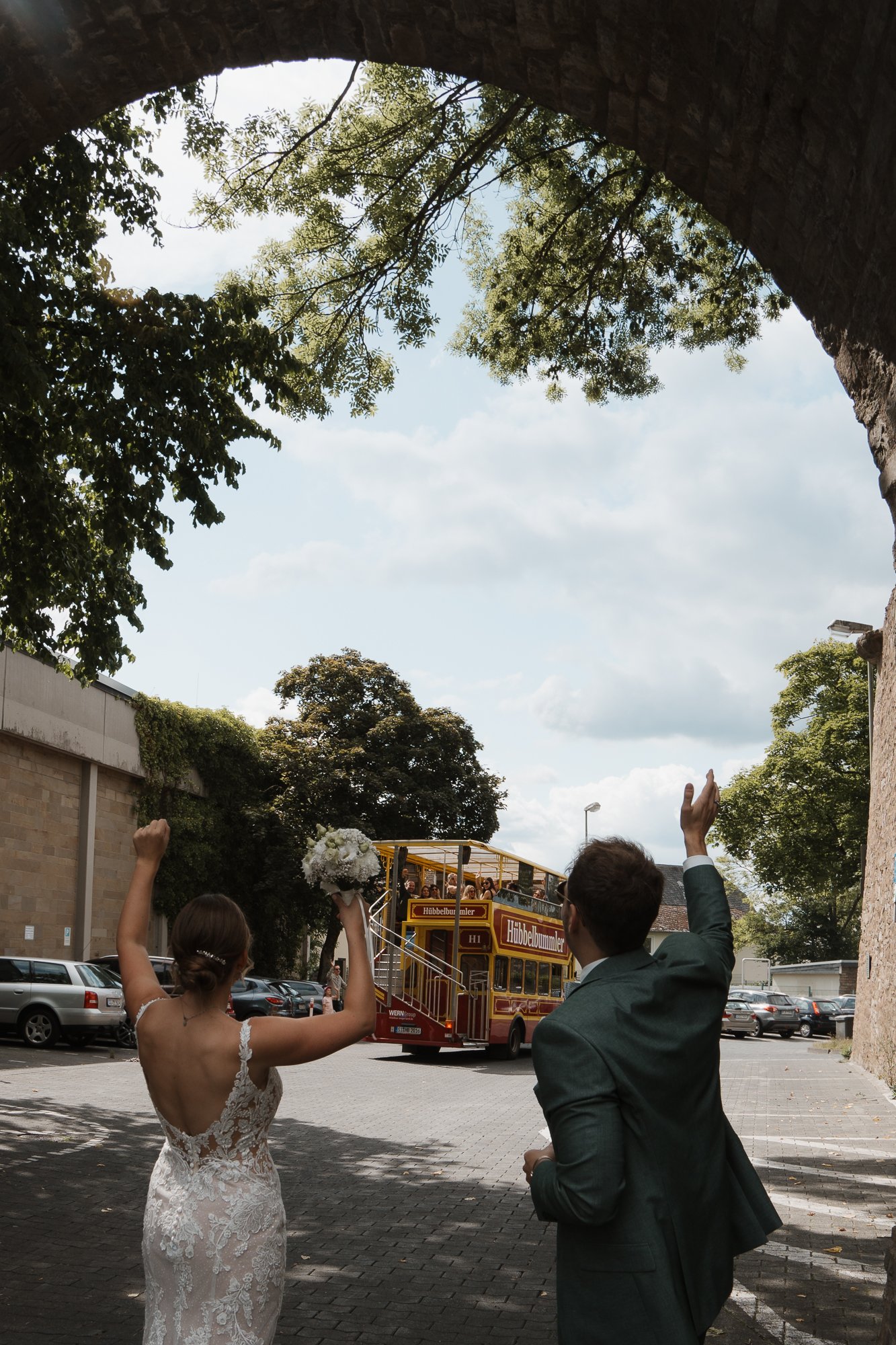 Ein Brautpaar feiert, die Braut hält einen Blumenstrauß, der Bräutigam winkt, im Hintergrund fährt eine Attraktion namens 'Hüpfebummmler' mit mehreren Personen. Es ist draußen bei Tageslicht, umgeben von Bäumen und Parkplätzen.