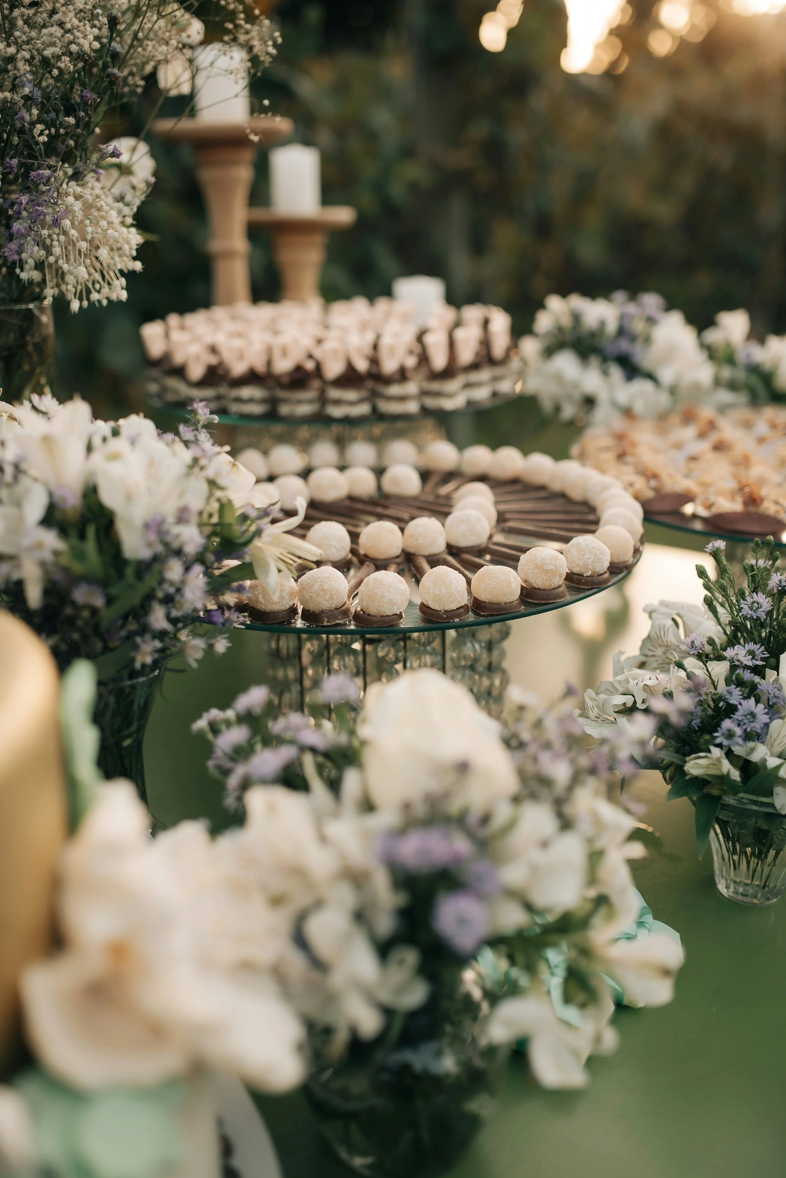 Dessert table with white and brown treats, surrounded by white floral arrangements, set outdoors during sunset.