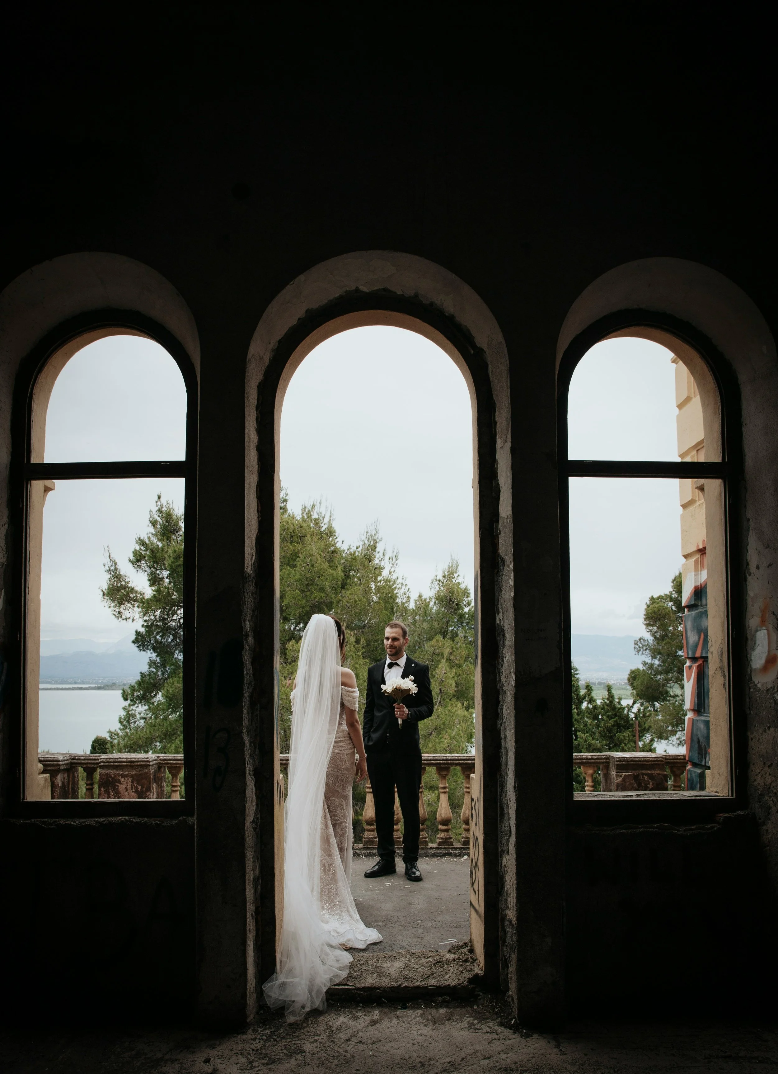 A bride and groom on a balcony, viewed through three arched windows from inside an abandoned building with graffiti, overlooking trees and a body of water in the background.